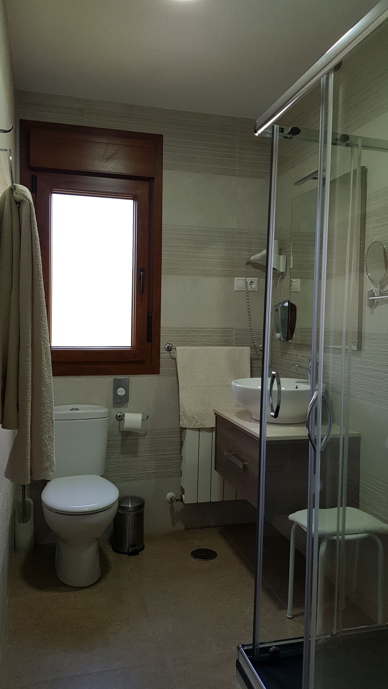 Baño con inodoro, lavabo, ducha y ventana. Paredes claras y marco de madera oscura.