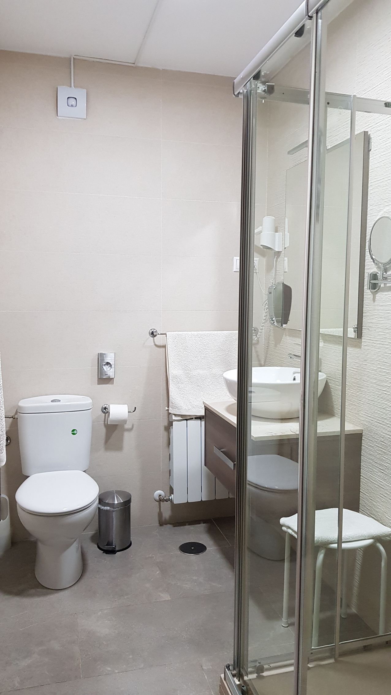 Baño con inodoro, ducha, tocador y un pequeño taburete. Paredes beige, suelo gris y mampara de cristal.