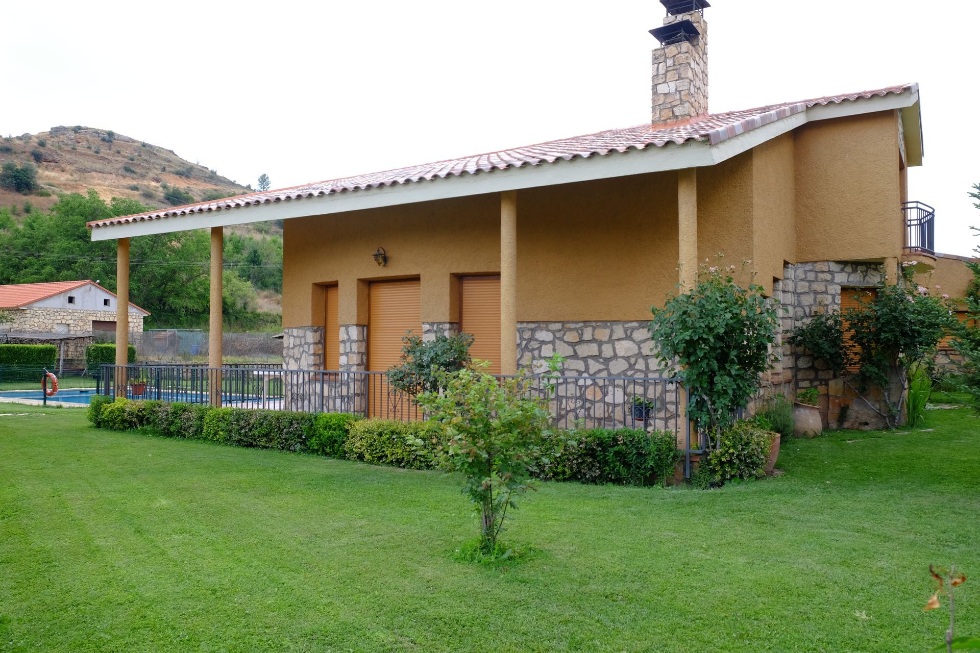 Casa de color tostado con detalles de piedra, techo de tejas rojas y césped verde en un entorno rural.