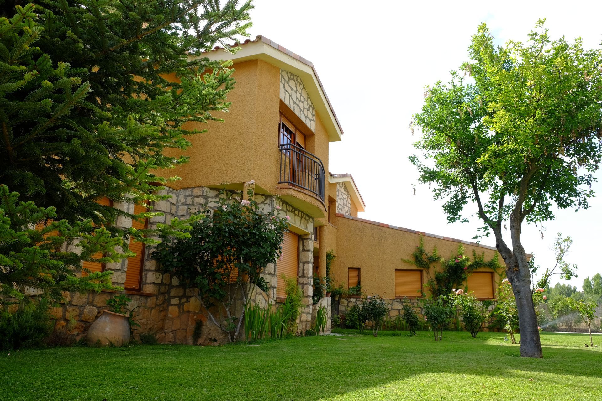 Casa con exterior de estuco color canela, base de piedra y balcón, rodeada de césped verde y árboles.