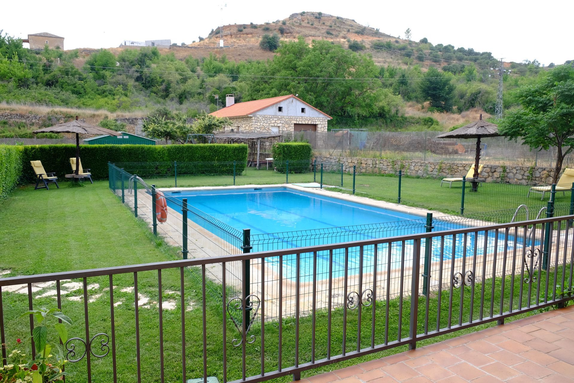 Piscina rodeada de césped verde y vallada, con un edificio y colina al fondo.