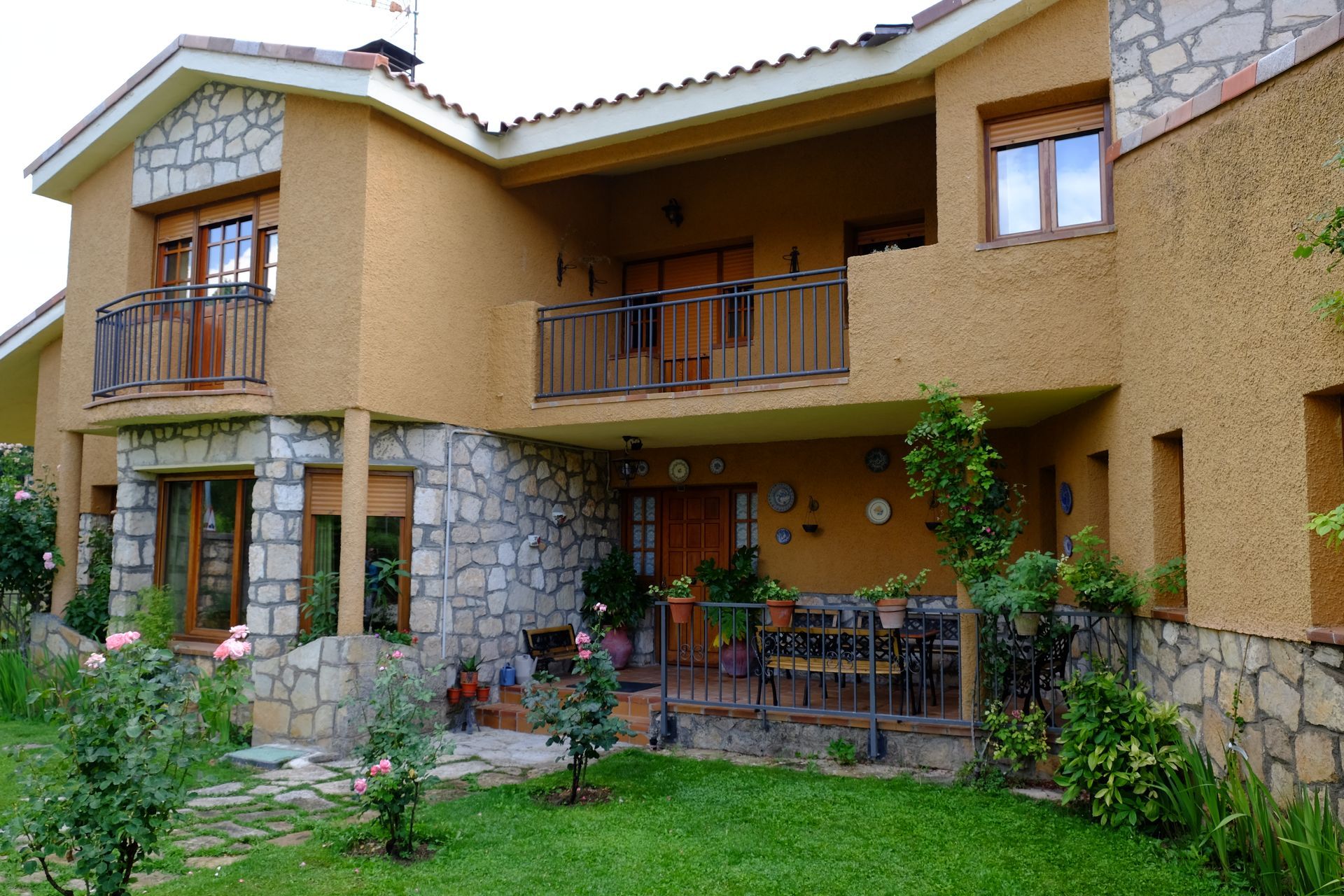 Casa de dos pisos con estuco color canela, detalles de piedra, balcones y un patio con césped y flores.