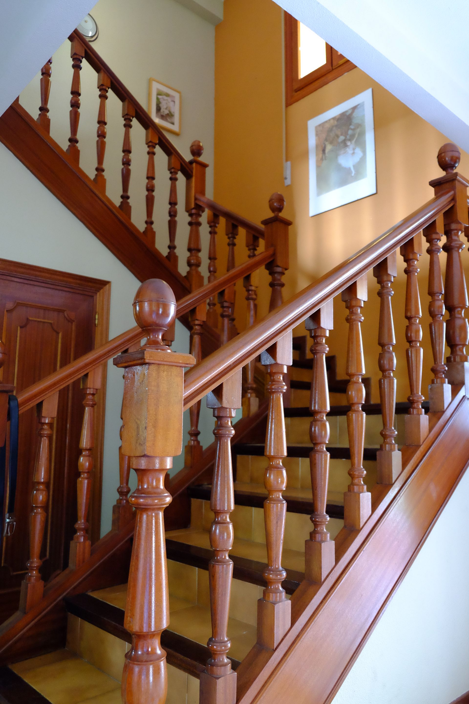 Escalera de madera con barandillas y pasamanos en una casa. La pared está pintada de un naranja cálido y hay una obra de arte colgada.