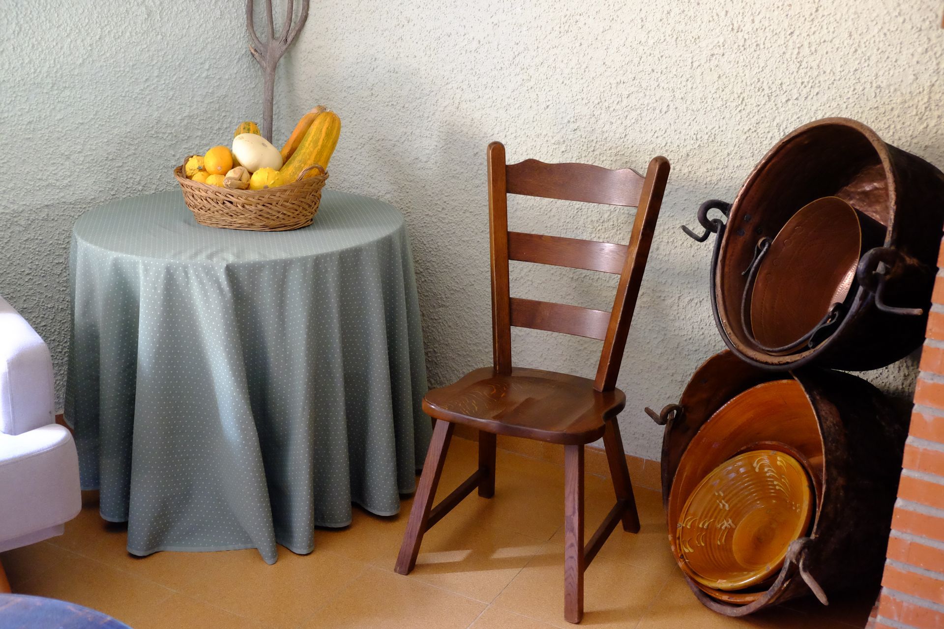 Un rincón de cocina rústico con una silla de madera, una mesa redonda con fruta y ollas de cobre apiladas.
