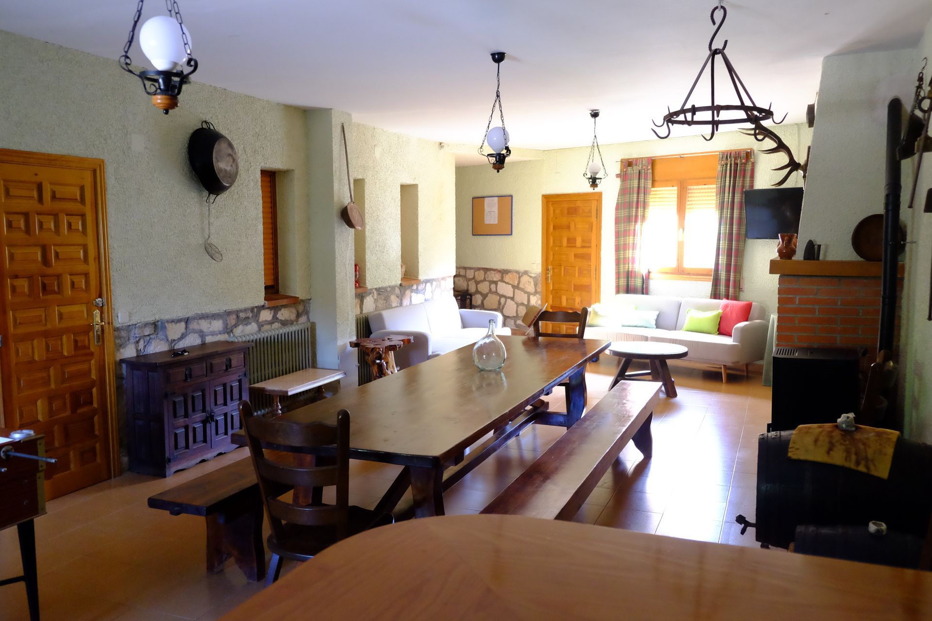 Espaciosa sala de estar rústica con una gran mesa y bancos de madera. Paredes beige, detalles en piedra y varias lámparas colgantes.