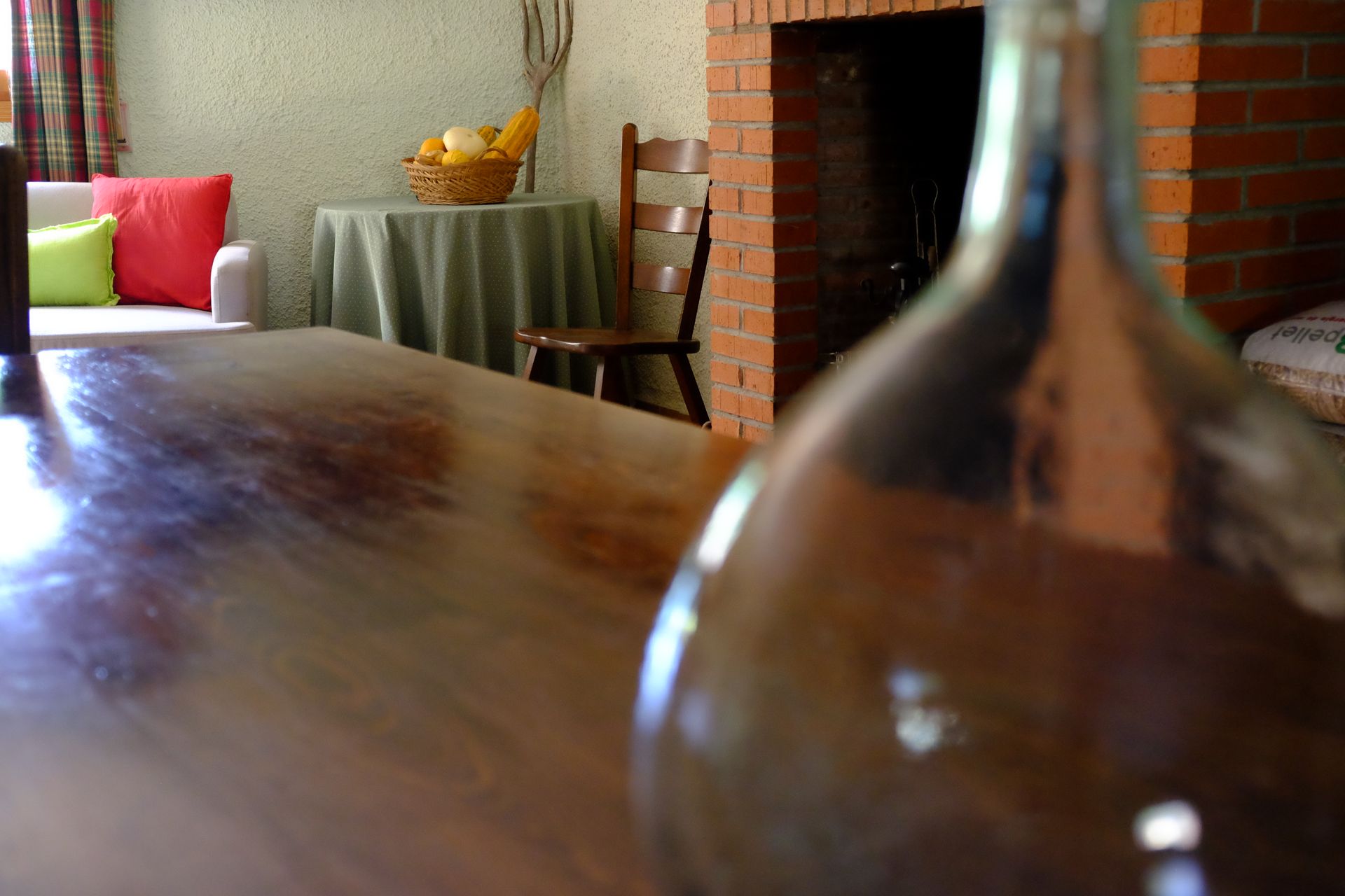 Mesa de madera en primer plano, botella de vidrio desenfocada. Fondo de la sala de estar: chimenea, silla, mesa con cesta de fruta y sofá.