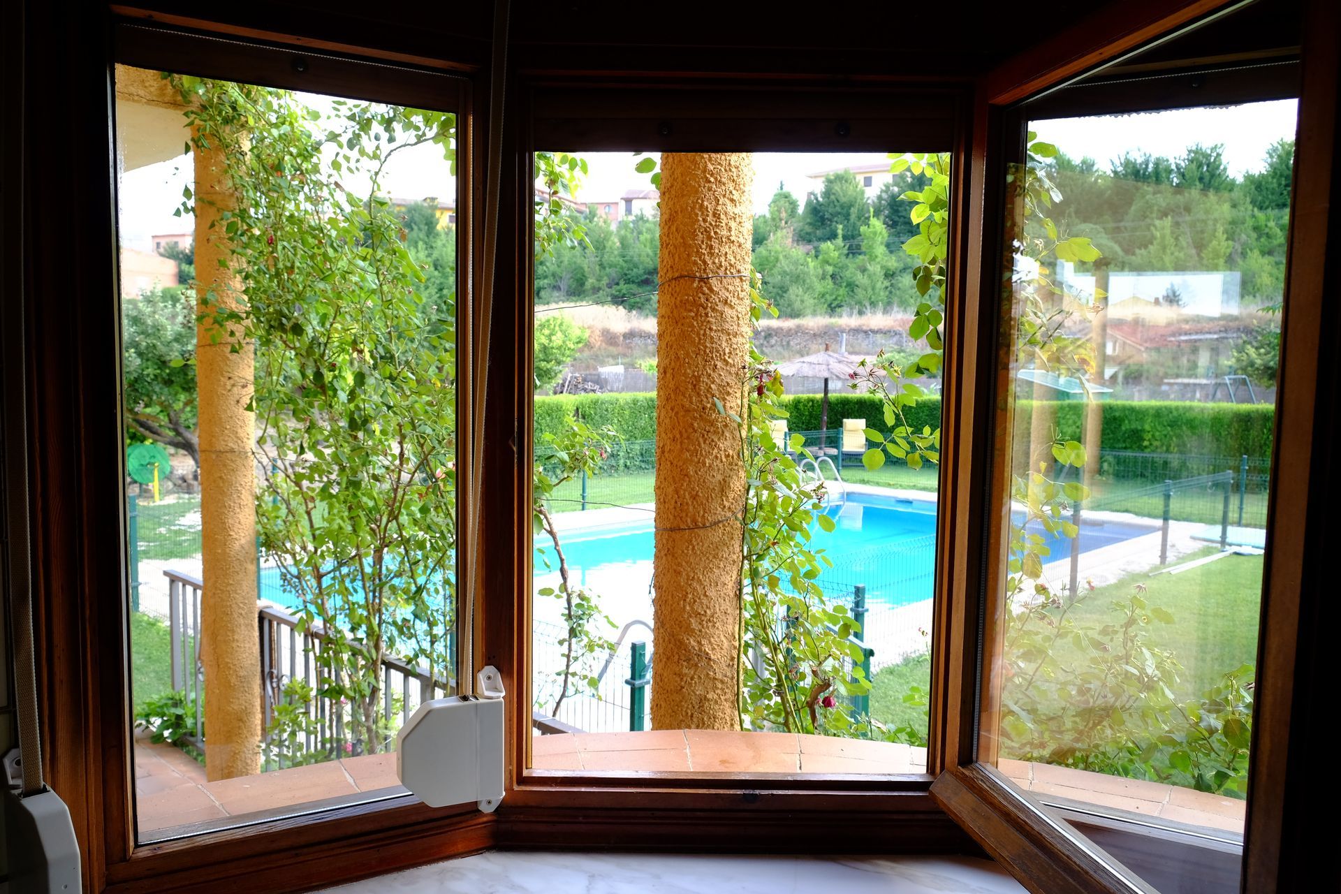 Ventanal con vistas a una piscina, con árboles y césped verde.
