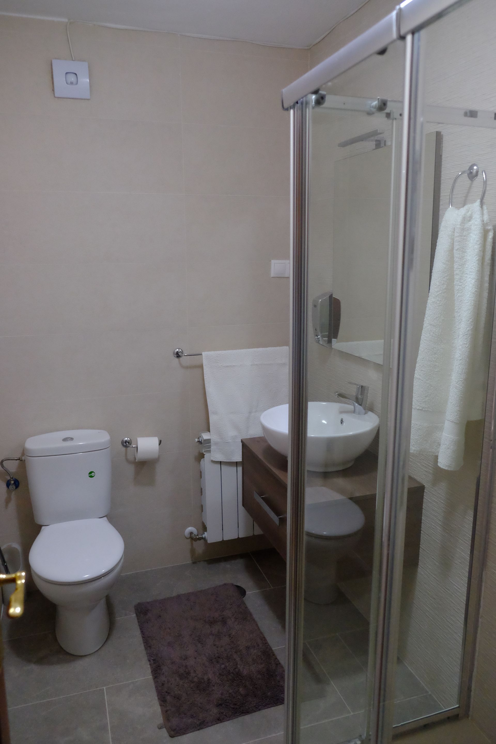 Baño con inodoro, lavabo y ducha con mampara de cristal. Paredes beige, grifería blanca y alfombra oscura.