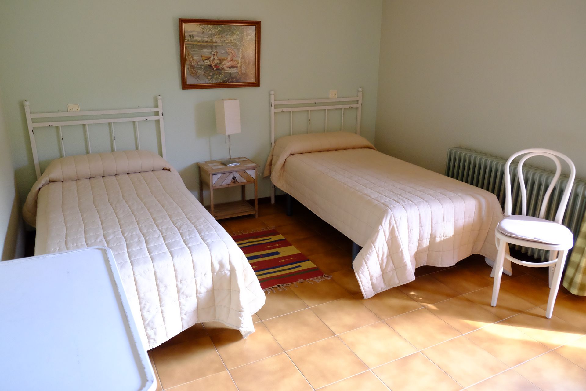 Dormitorio con dos camas blancas, mesita de noche, silla, radiador y una pequeña alfombra sobre suelo de baldosas.