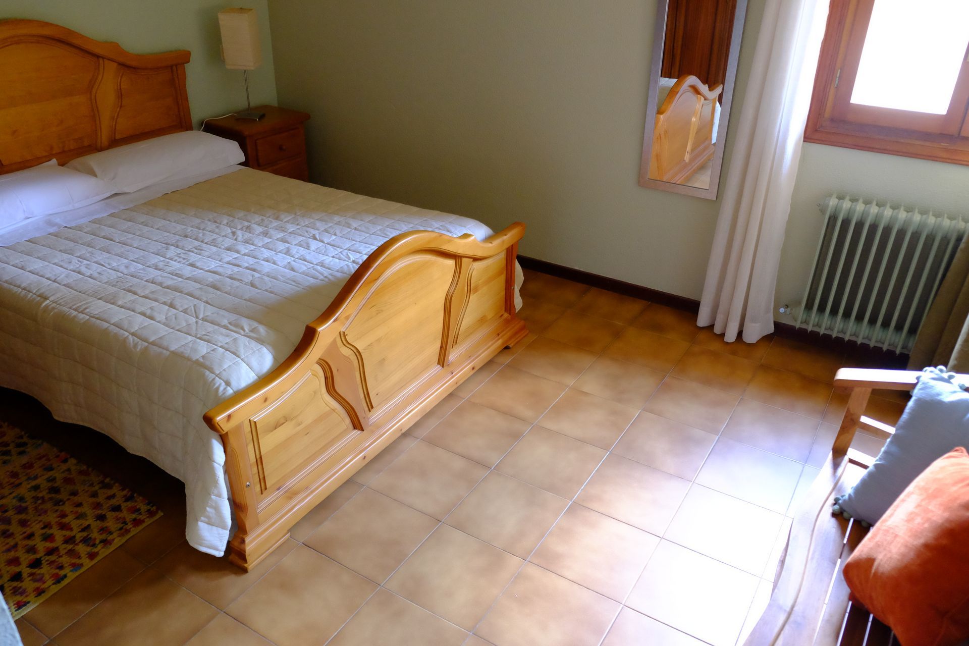 Dormitorio con cama de madera clara, mesita de noche y suelo de baldosas. También se ven una silla y una ventana.