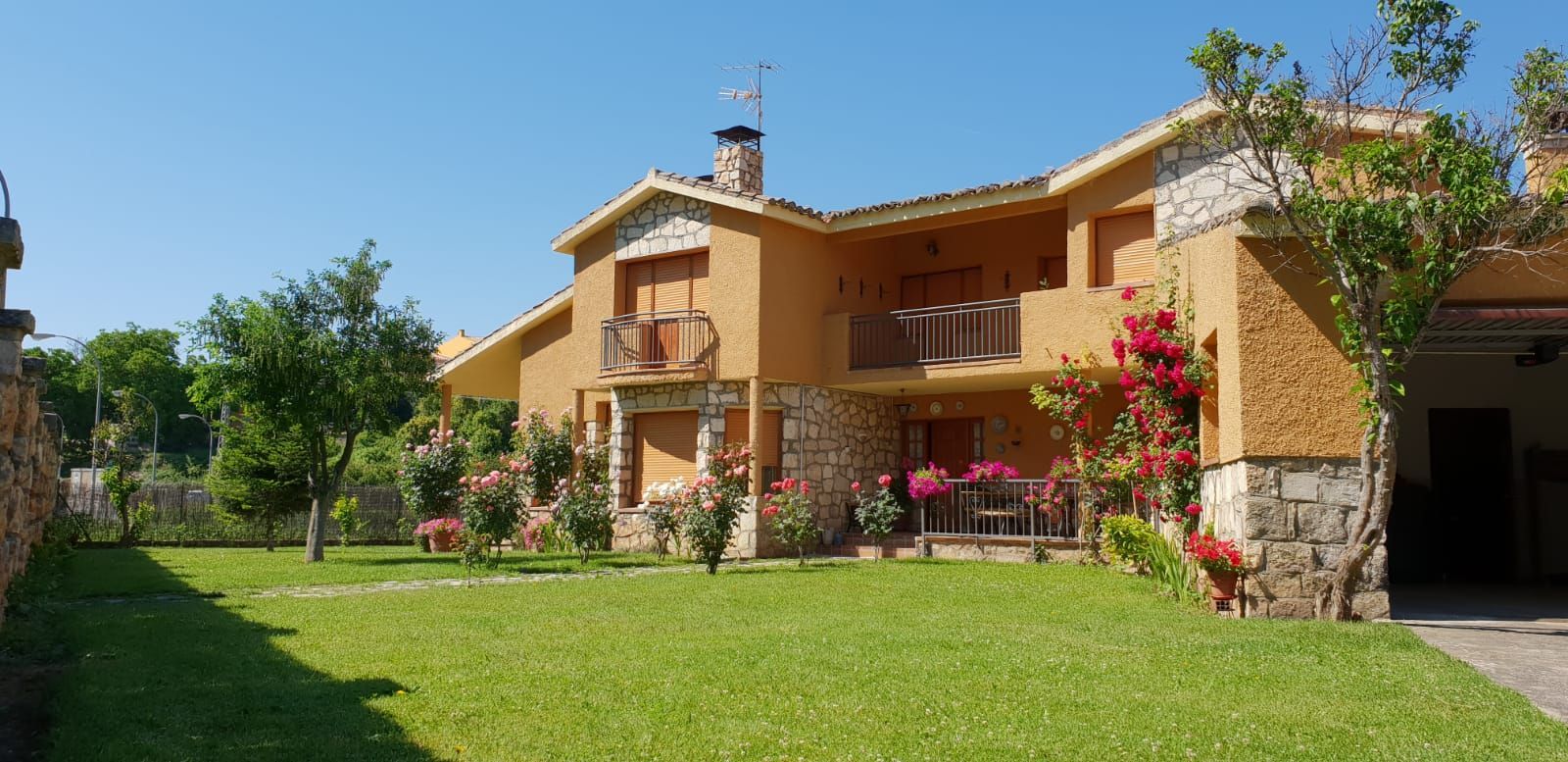 Una casa de dos pisos con fachada de piedra y color canela, césped verde y flores bajo un cielo azul.