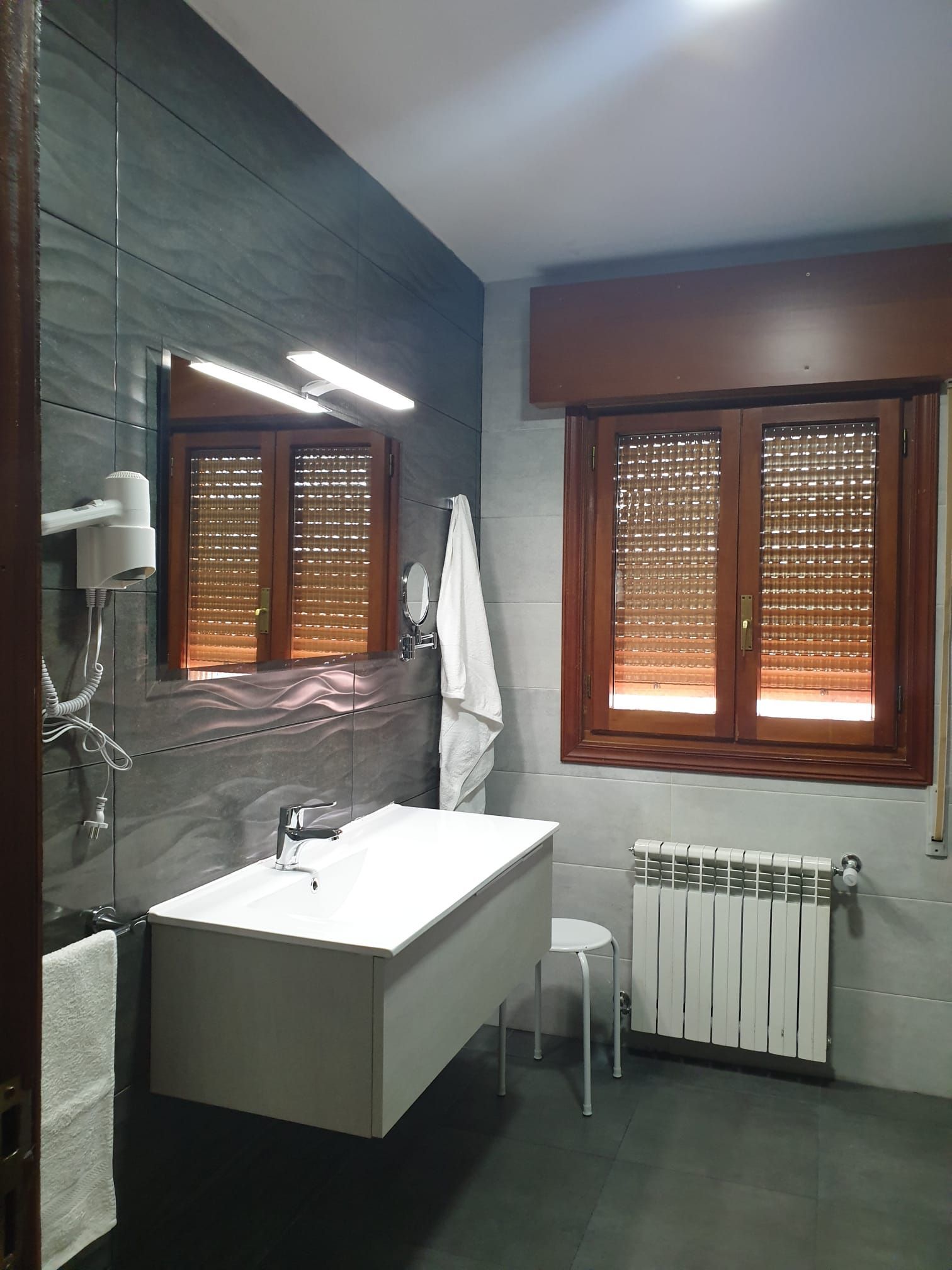 Baño con paredes de azulejos grises, lavabo flotante blanco, ventanas con contraventanas marrones y un radiador blanco.