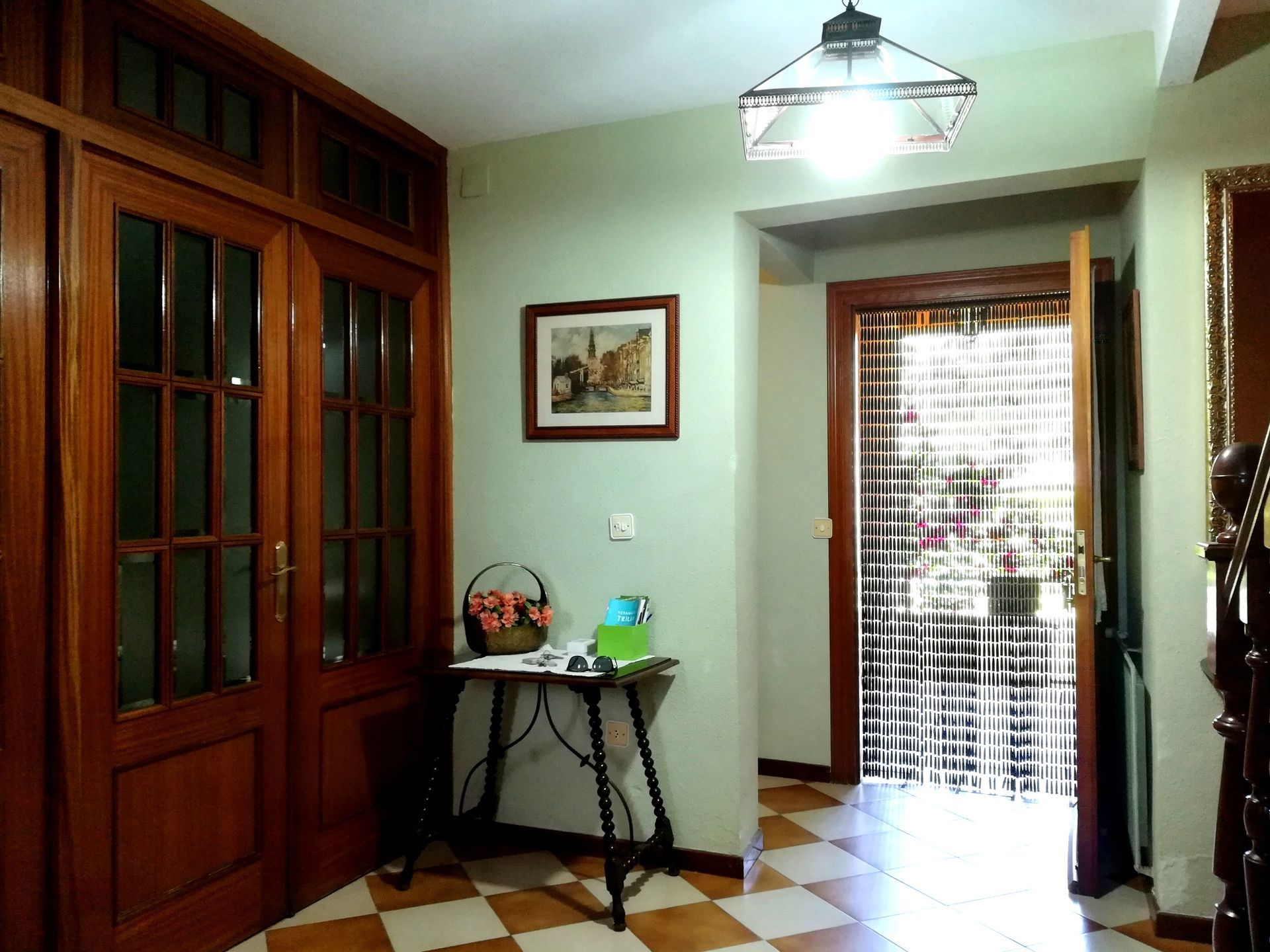 Pasillo con puertas de madera, suelo ajedrezado y puerta abierta que da al exterior.