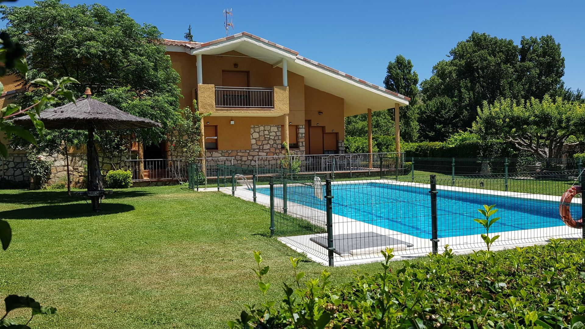 Casa con piscina en un patio con césped bajo un cielo azul; una sombrilla da sombra al césped.