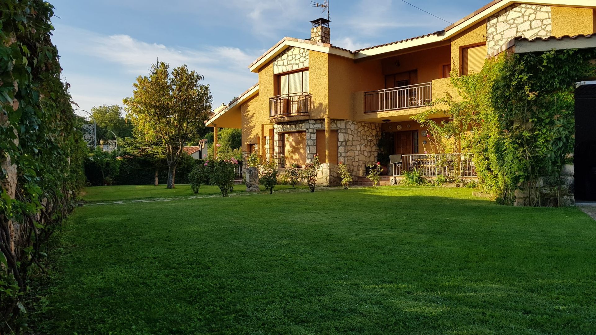 Casa de dos plantas con detalles en piedra, balcón, chimenea y amplio césped. La hiedra verde cubre las paredes, un día soleado.