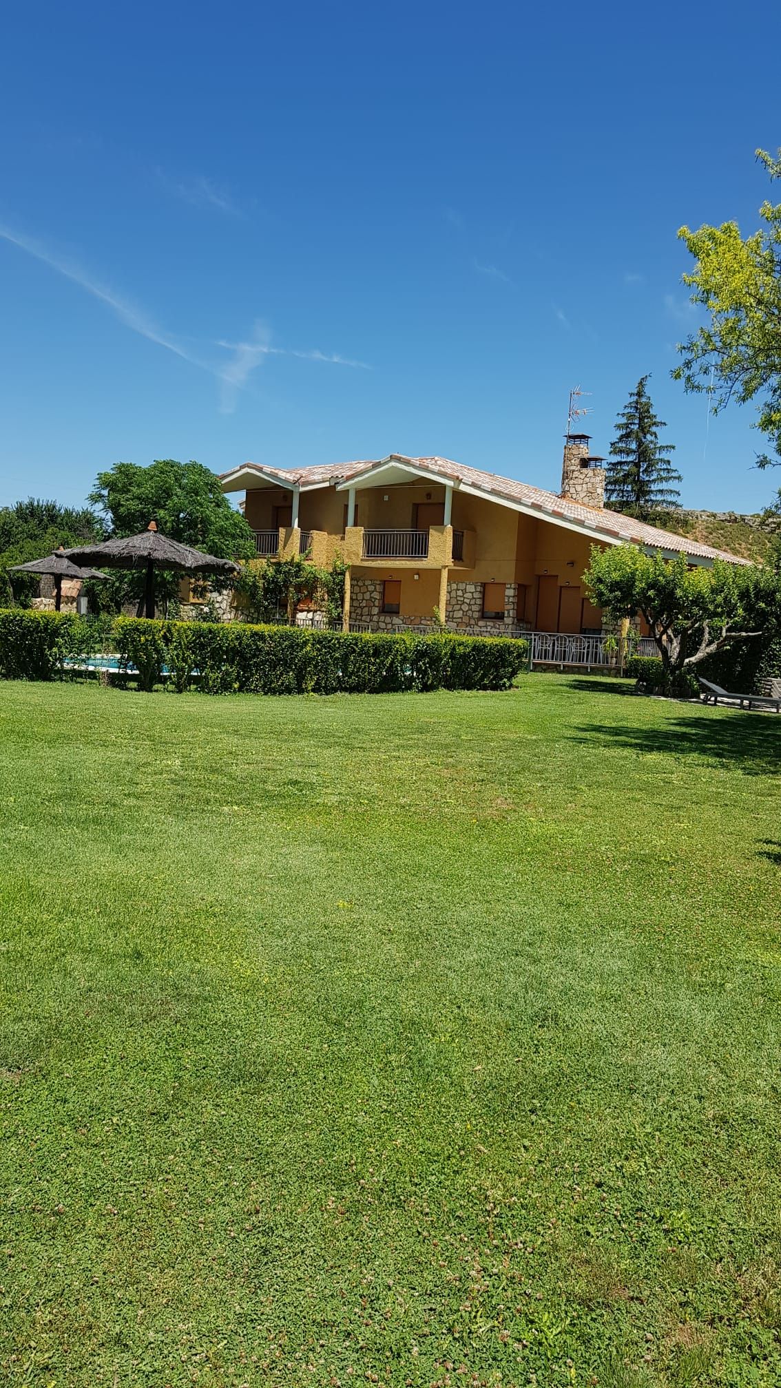 Casa amarilla de dos pisos con balcón, ubicada en un césped verde con árboles y un cielo azul claro.