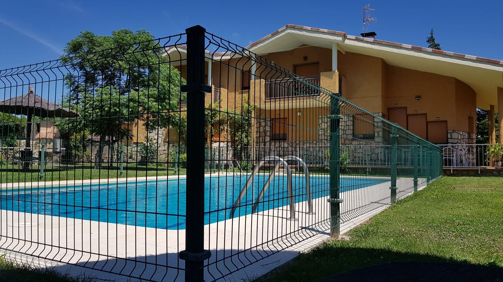 Una piscina vallada frente a una casa de dos pisos de color bronce con césped verde y árboles bajo un cielo soleado.