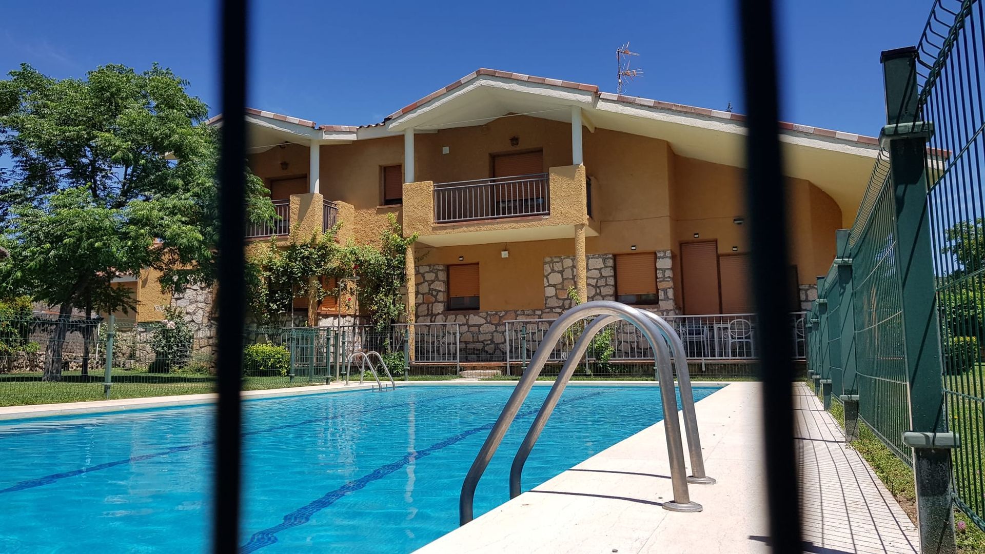 Piscina frente a casa de dos pisos con balcón, vista a través de una reja.