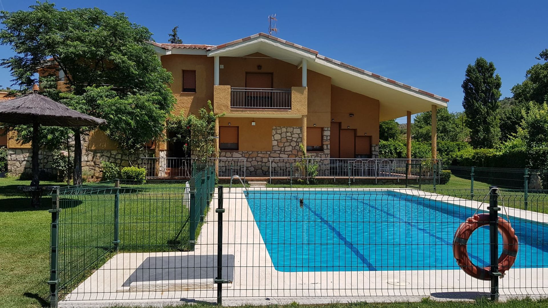 Casa de dos pisos con piscina tras una valla negra en un día soleado. Un salvavidas cuelga de la valla.