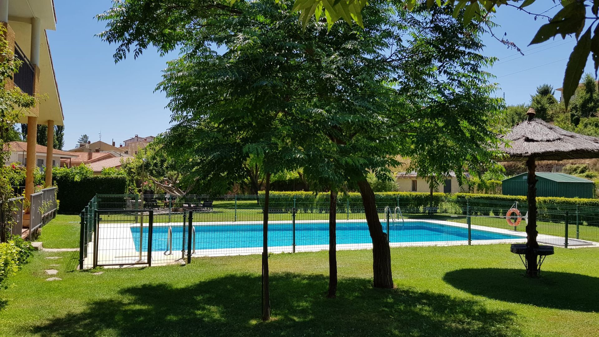 Piscina y césped frente a edificios de apartamentos bajo un cielo soleado, con árboles y una sombrilla de paja.