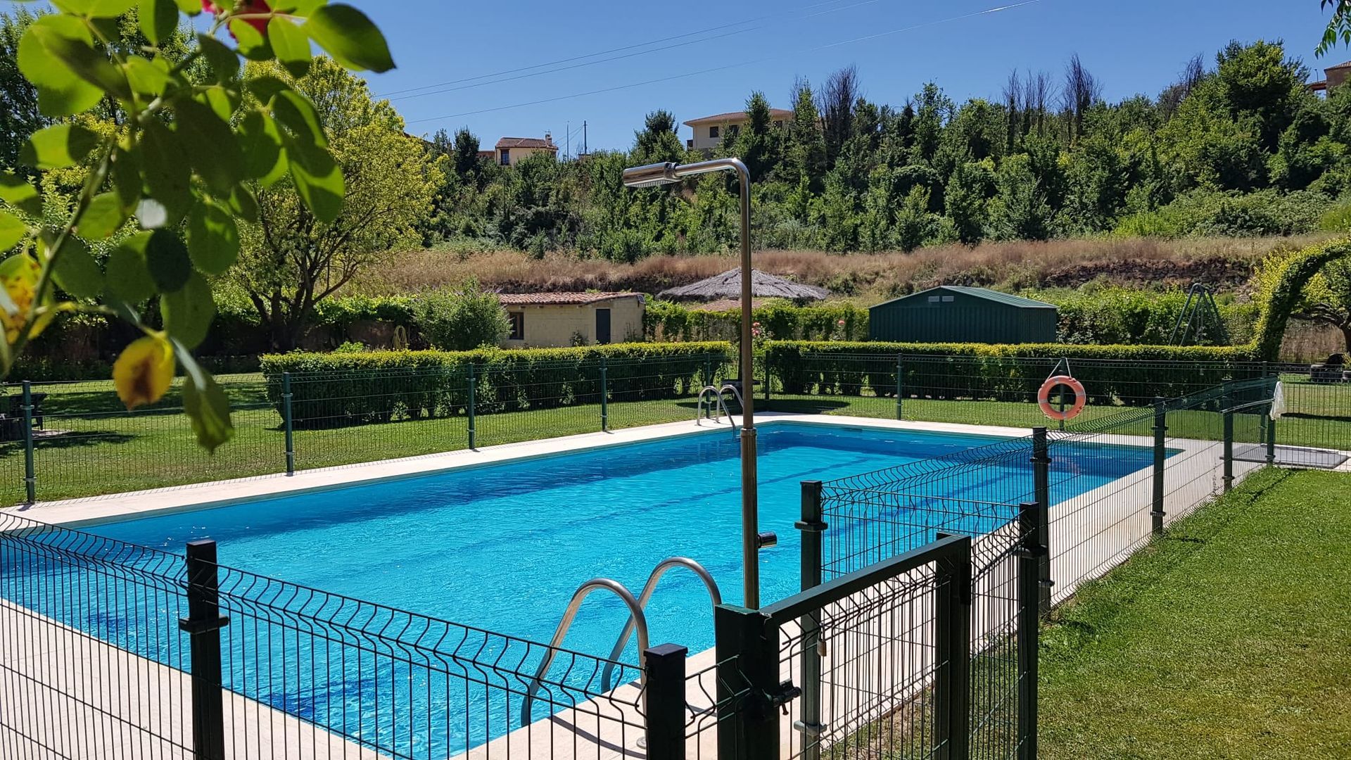 Piscina rodeada de césped verde, valla negra y árboles bajo un cielo soleado.