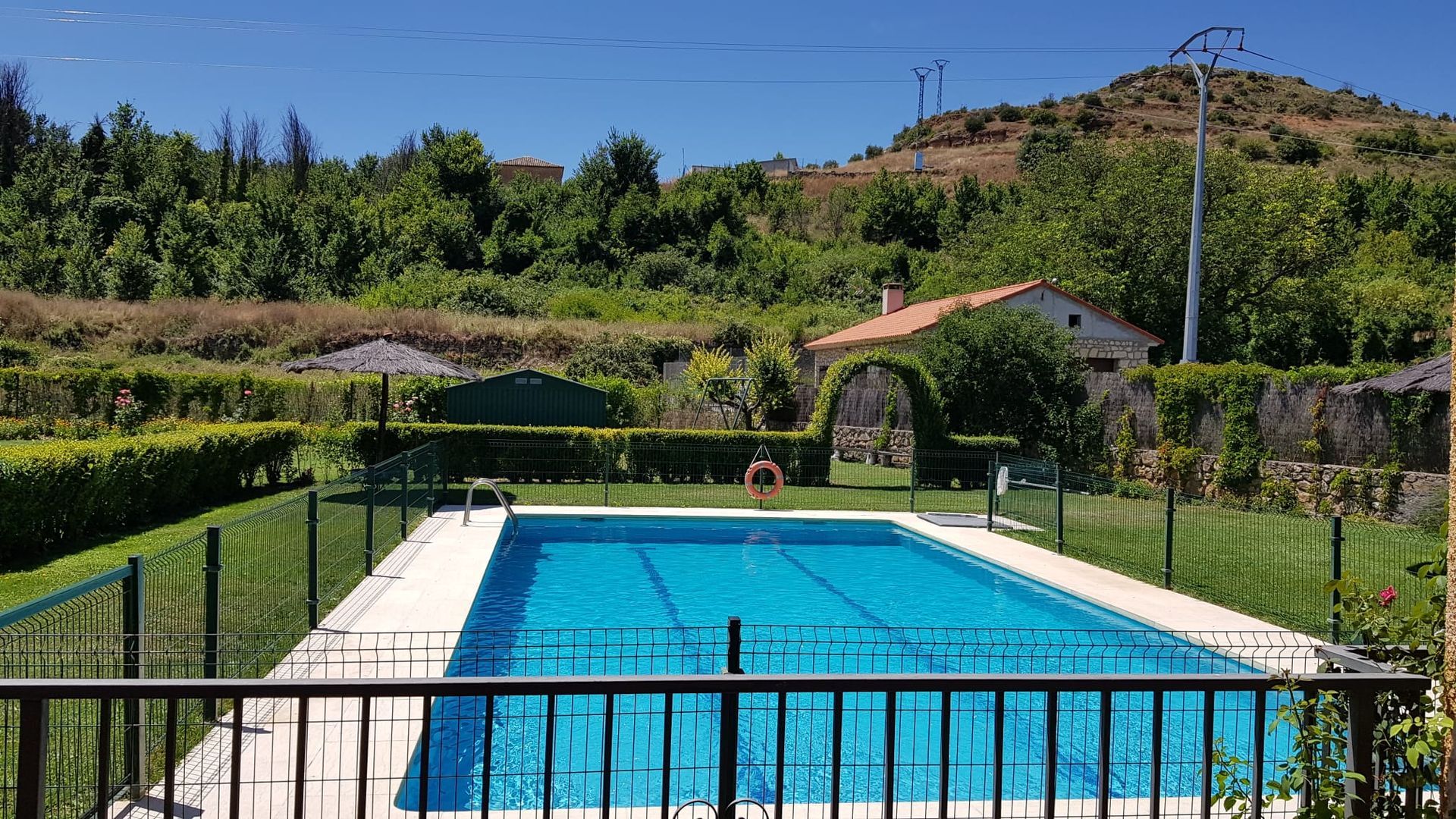 Piscina rodeada de setos verdes y un edificio de piedra bajo un cielo azul.