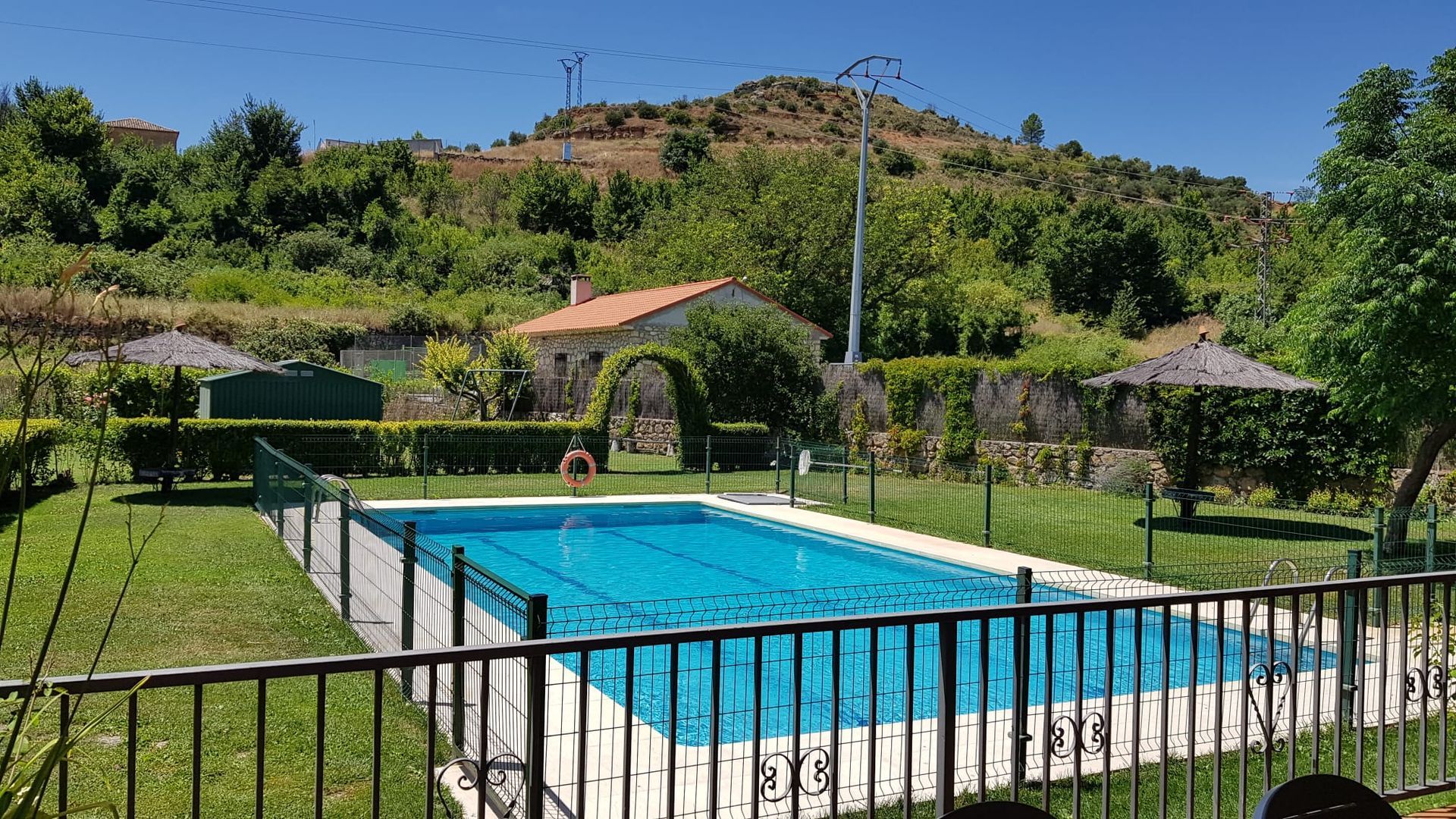 Piscina rodeada de césped y valla, con un edificio al fondo. Día soleado, montaña.