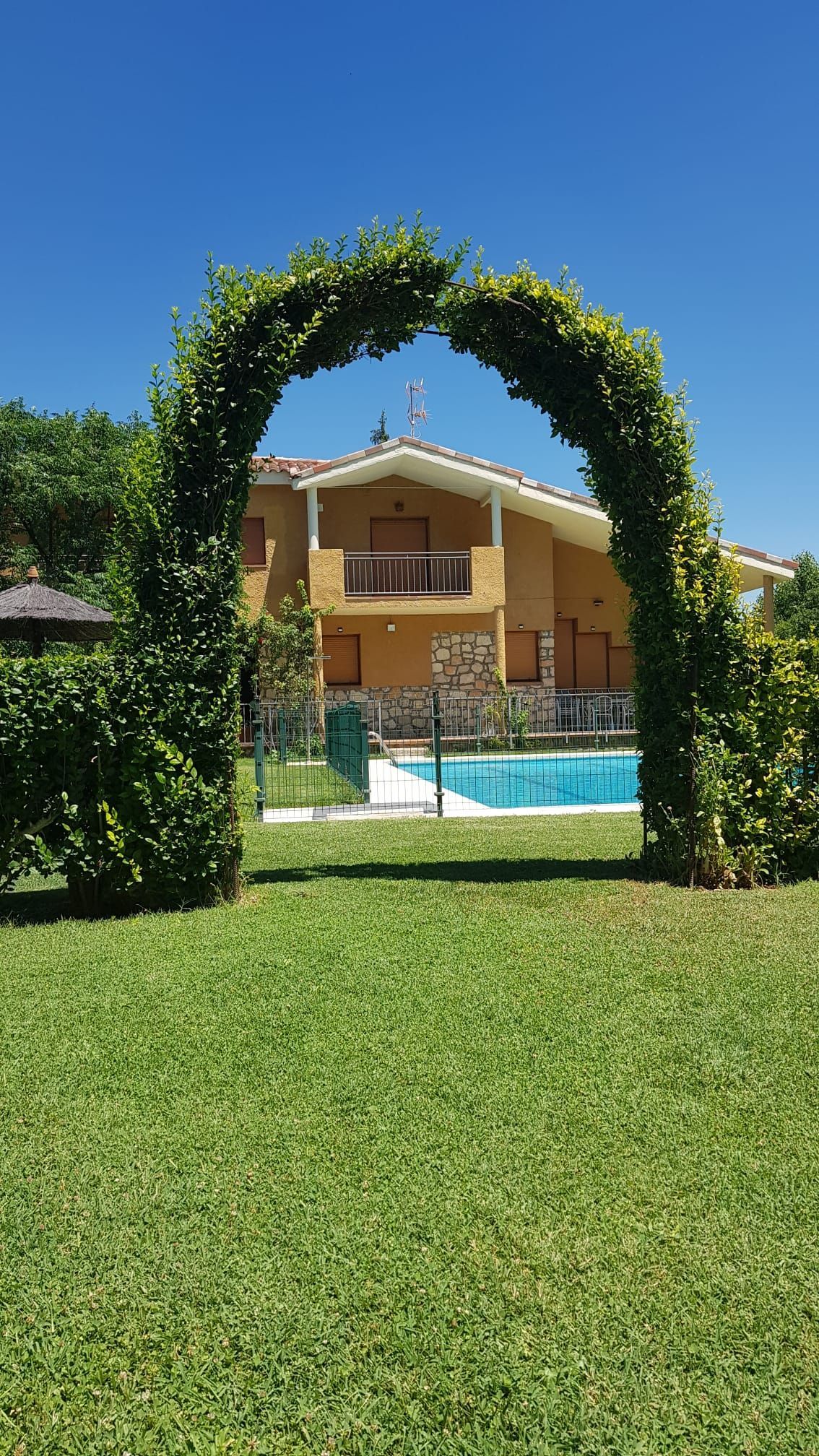 Un arco verde enmarca una vista de una casa amarilla con un balcón y una piscina bajo un cielo azul brillante.