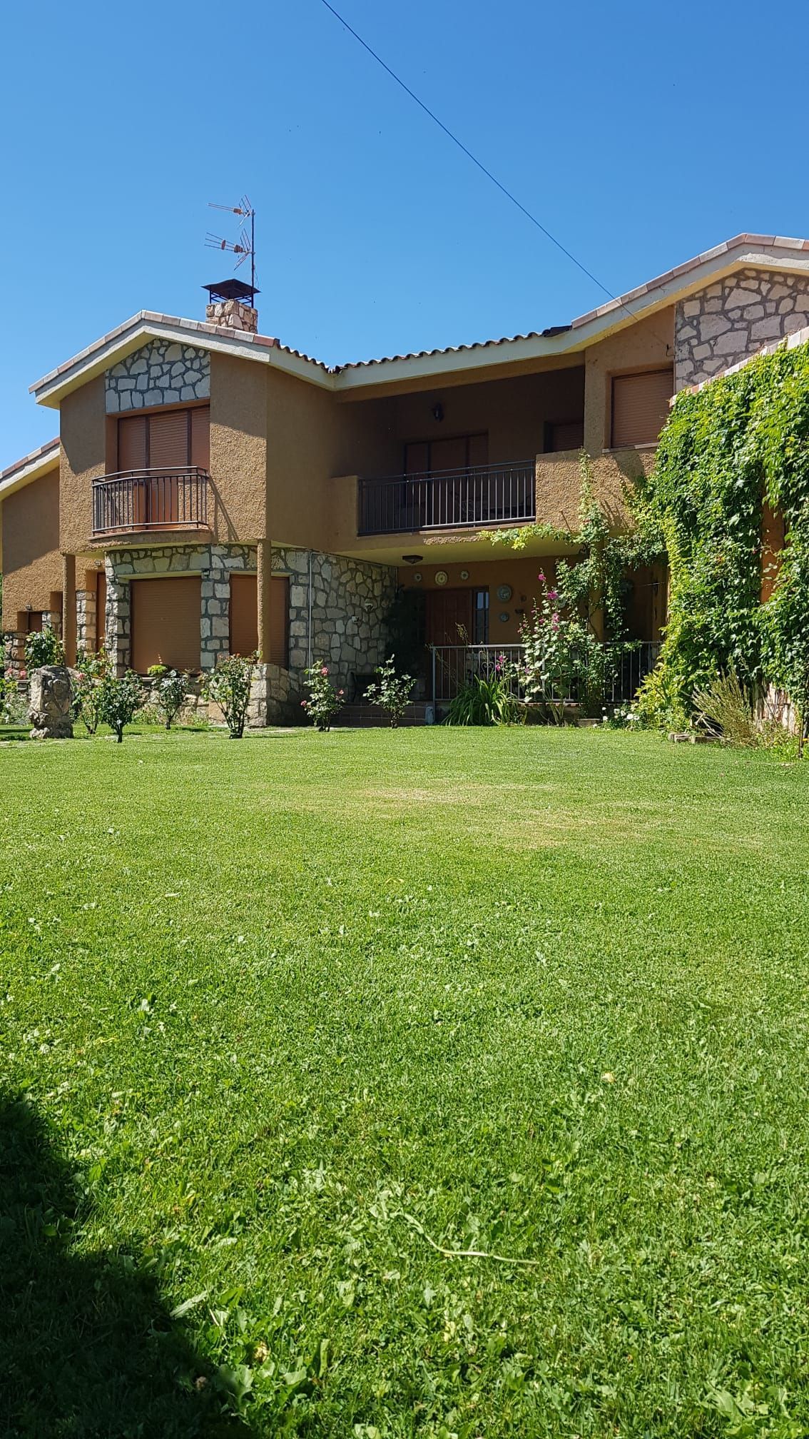 Casa de piedra de dos plantas con césped verde. Contraventanas de madera, balcones y plantas trepadoras. Cielo azul brillante.