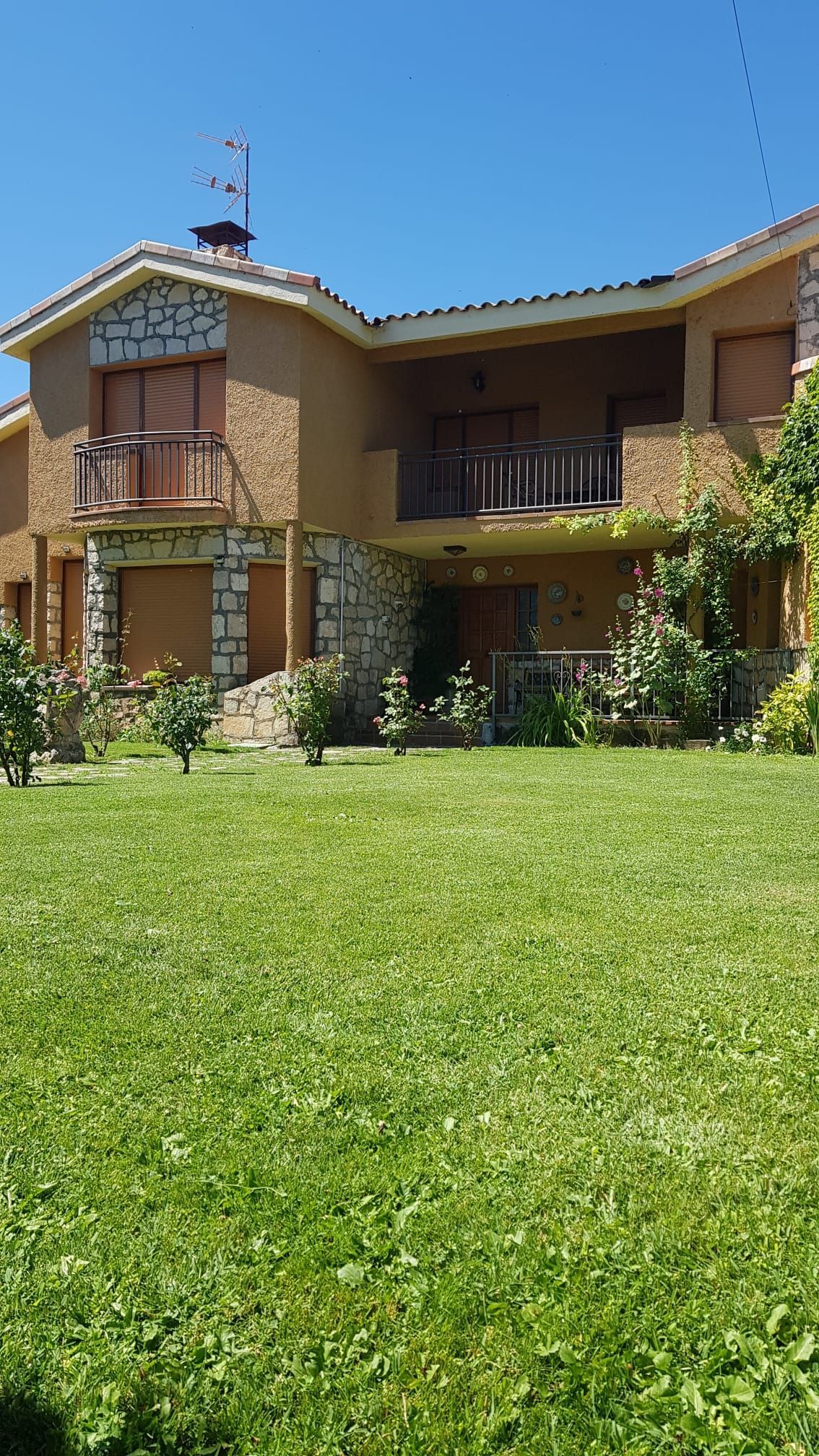 Casa de dos plantas con paredes color canela, detalles en piedra y balcones. Césped verde al frente bajo un cielo azul.
