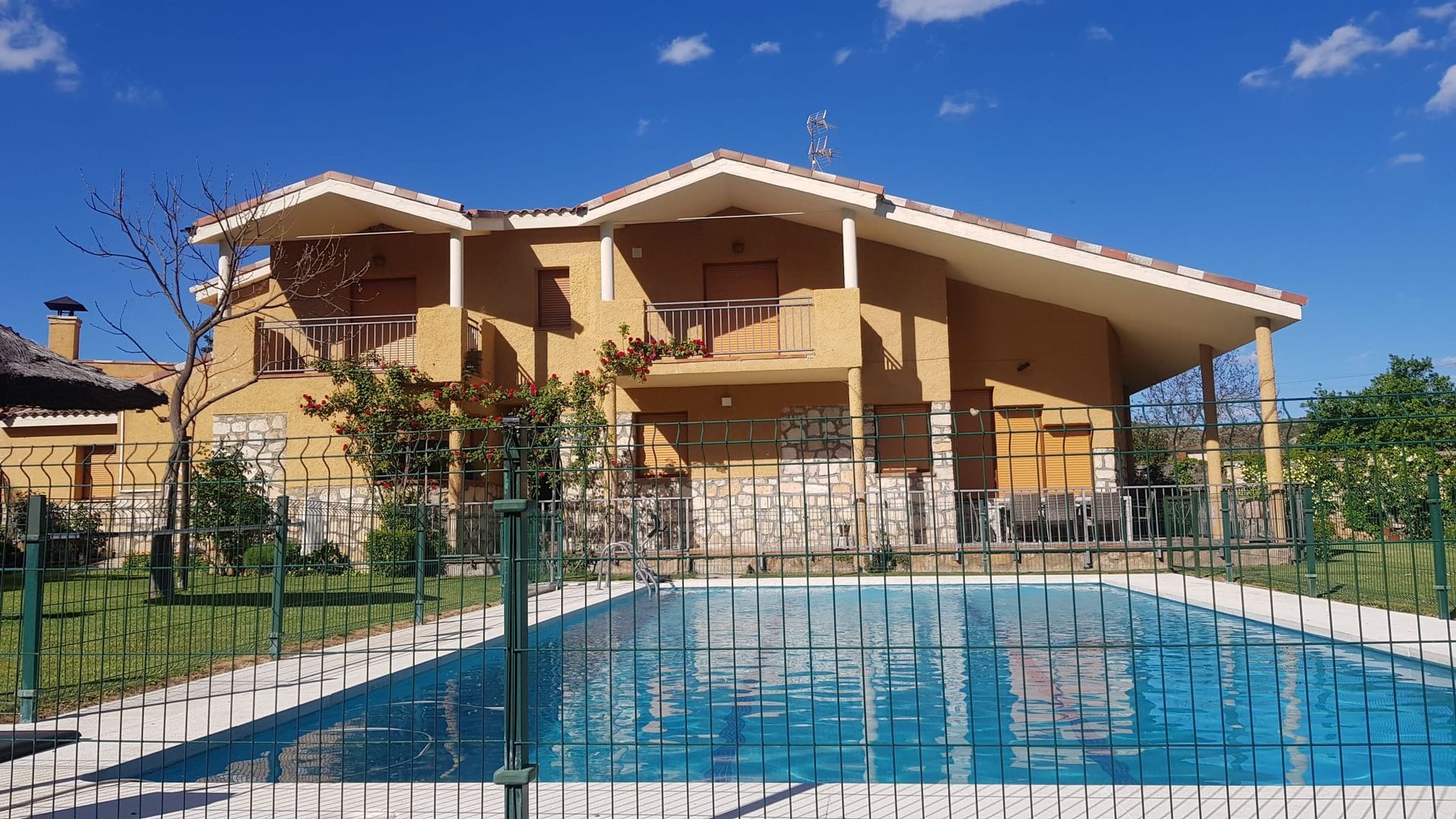 Una casa de dos pisos con piscina al frente, rodeada de una cerca verde, bajo un cielo azul.