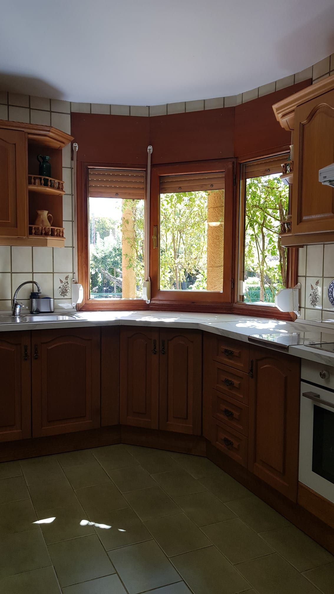 Cocina con gabinetes de madera, ventana curva con vista a la vegetación y encimera blanca.