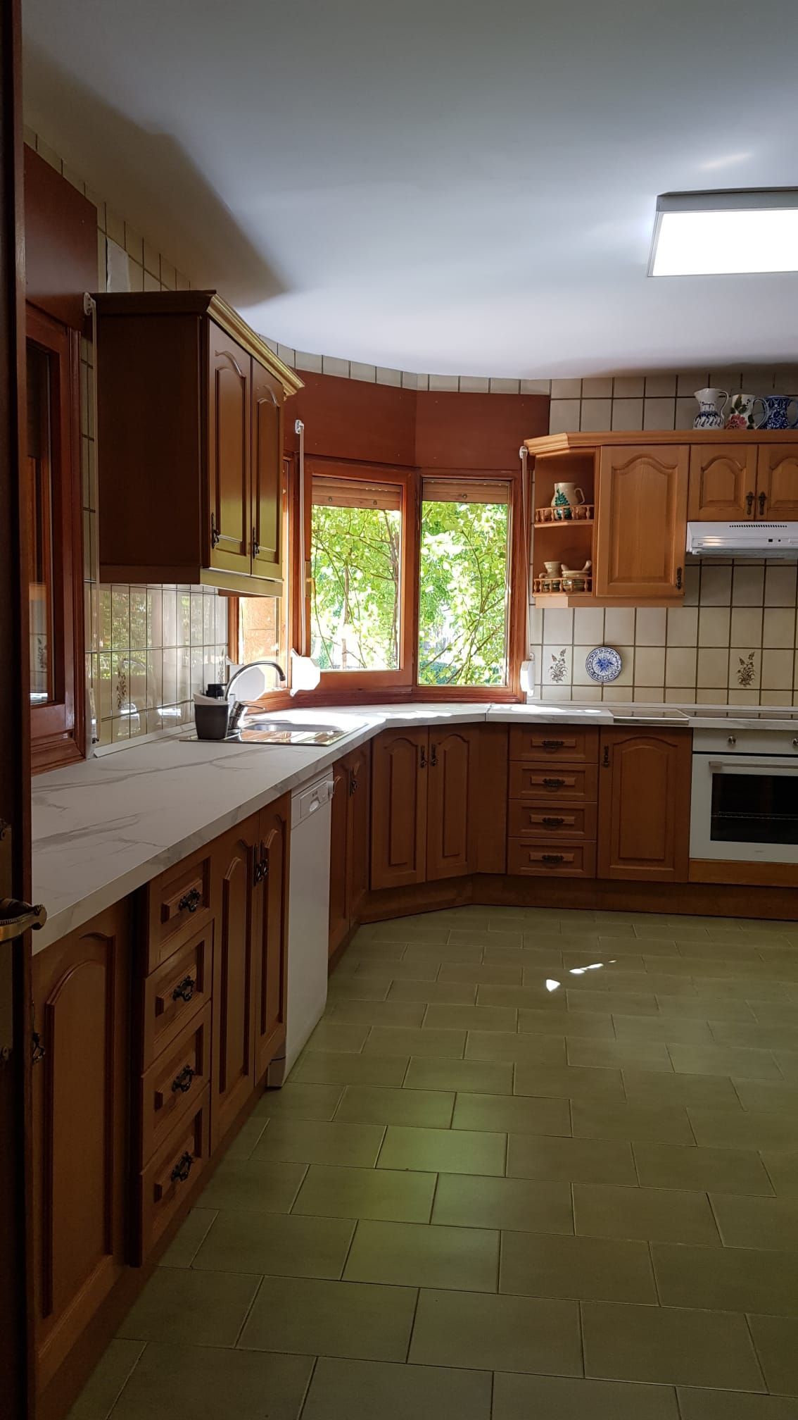 Cocina con gabinetes de madera, encimeras y una ventana con vista a la vegetación. Suelo verde y salpicadero de azulejos en tonos neutros.