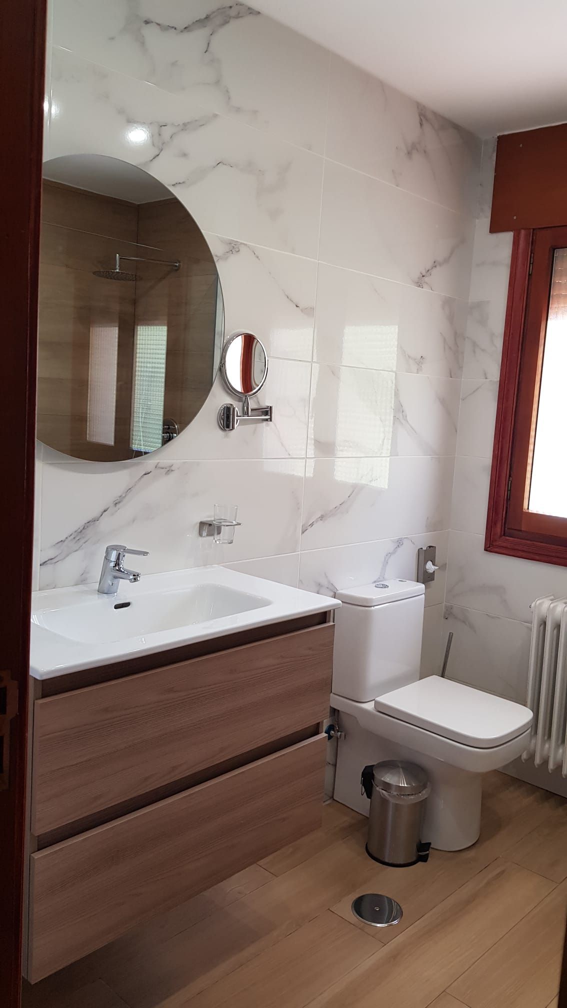 Baño con azulejos efecto mármol, tocador de madera, espejo redondo, inodoro blanco y ventana con marco de madera.