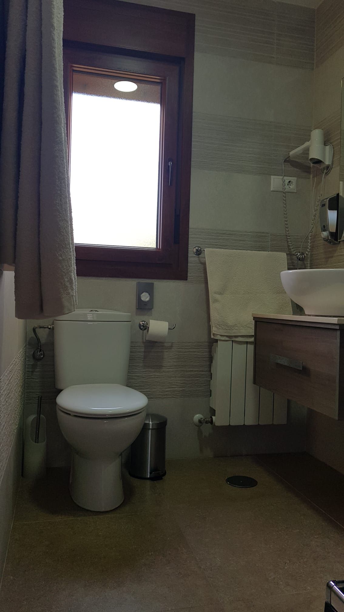 Baño con inodoro, ventana y lavabo. Tonos beige y marrón; toalla sobre el radiador.