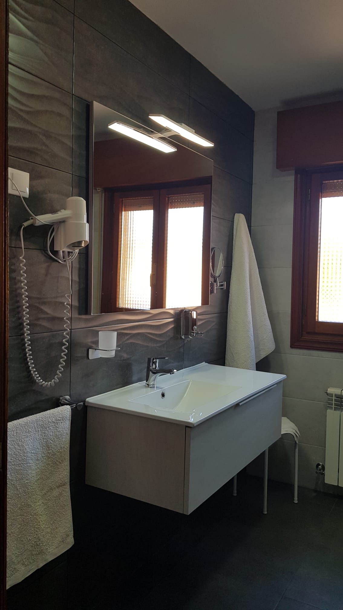 Baño con lavabo, espejo, toalla y ventana. Paredes de azulejos gris oscuro y suelo oscuro.