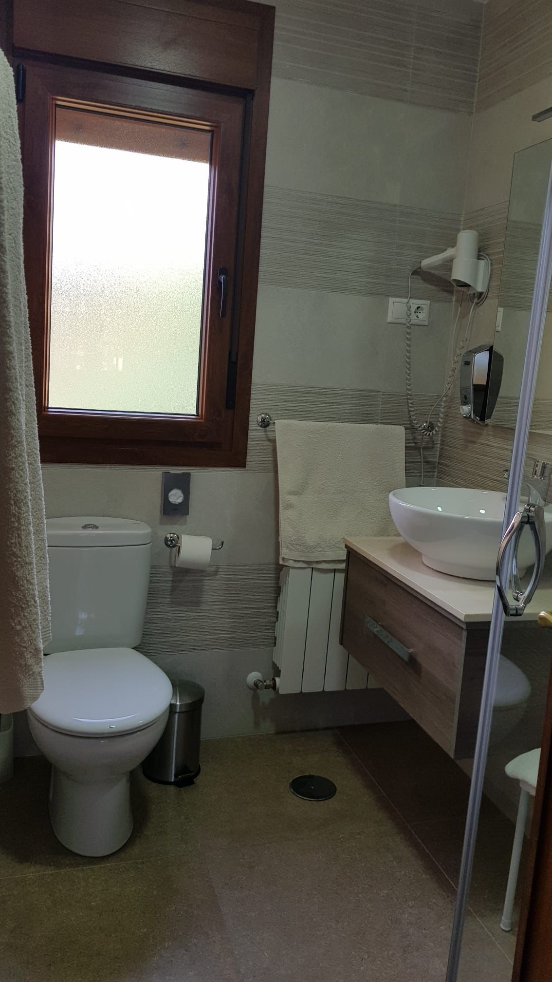 Baño con inodoro, lavabo y ventana. Paredes de azulejos beige. Molduras de madera marrón.