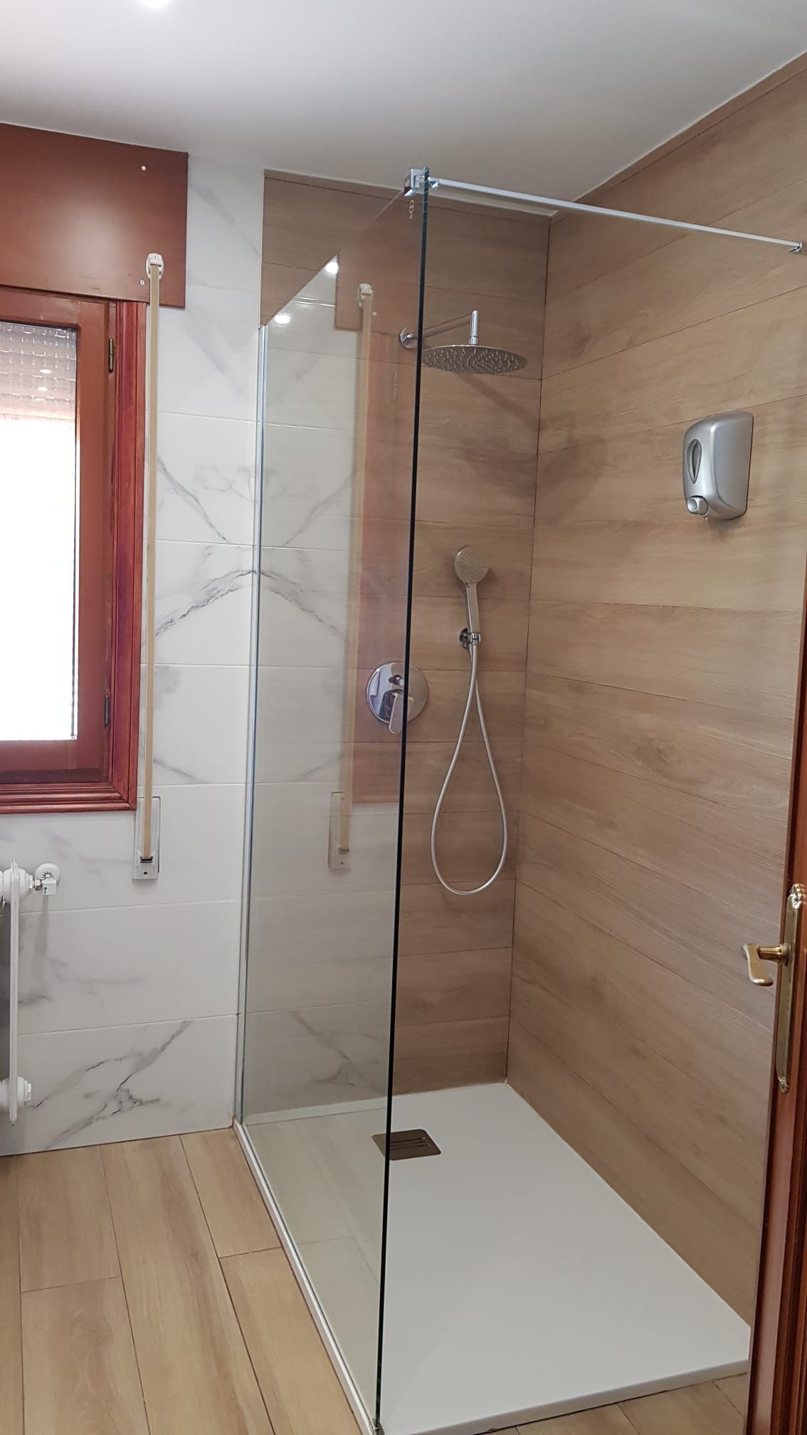 Cabina de ducha con puerta de cristal y azulejos imitación madera y mármol. Grifería plateada y base de ducha blanca.
