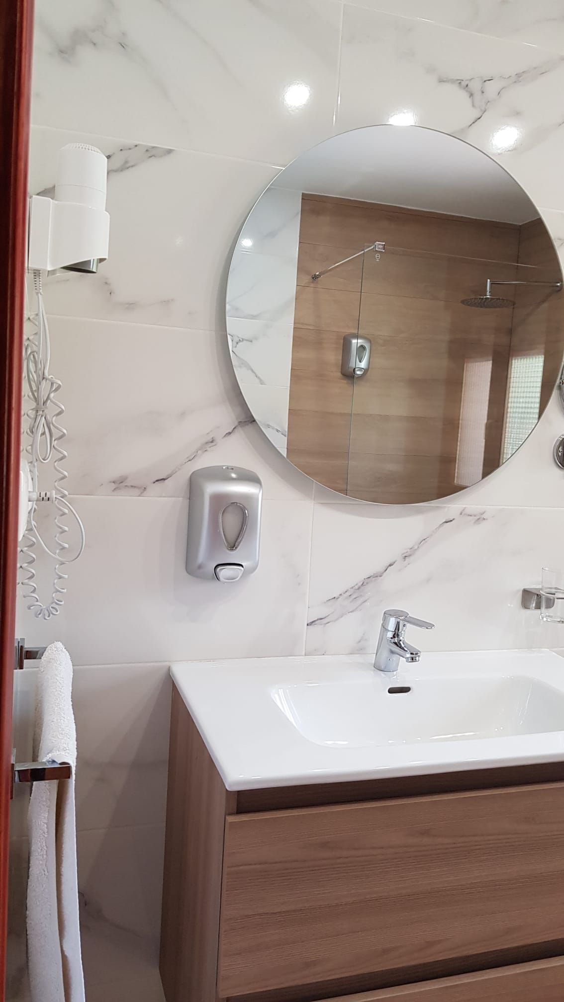 Baño con espejo redondo, lavabo blanco, mueble de madera y secador de manos.