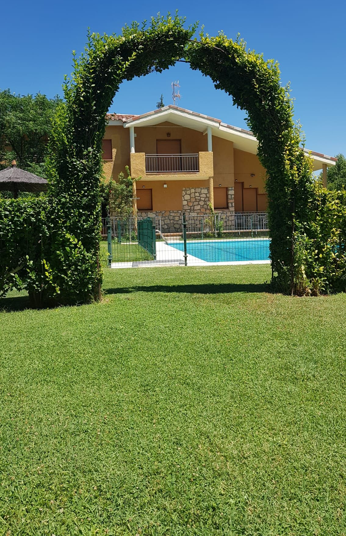 Una casa con piscina enmarcada por un arco verde, situada en un césped verde bajo un cielo azul.