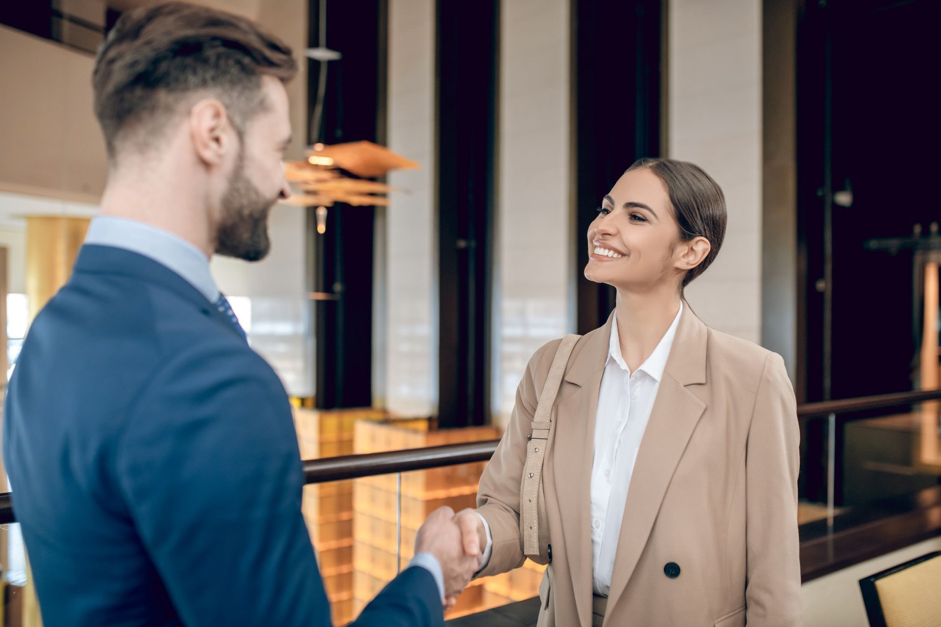 Un hombre con traje azul estrecha la mano a una mujer con chaqueta color canela, sonriendo en un vestíbulo.