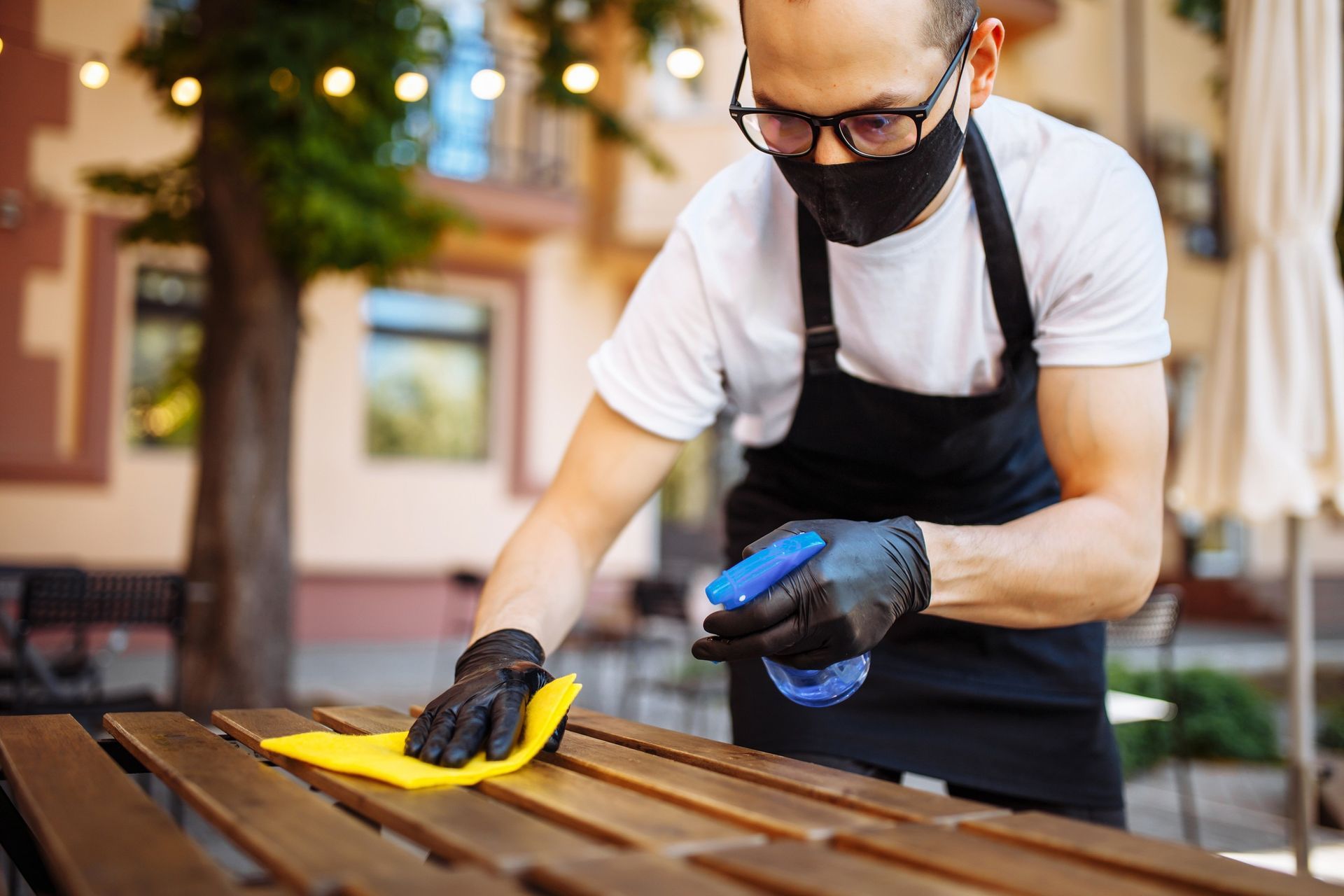 Persona con mascarilla y guantes desinfectando una mesa de madera al aire libre con un atomizador y un paño.