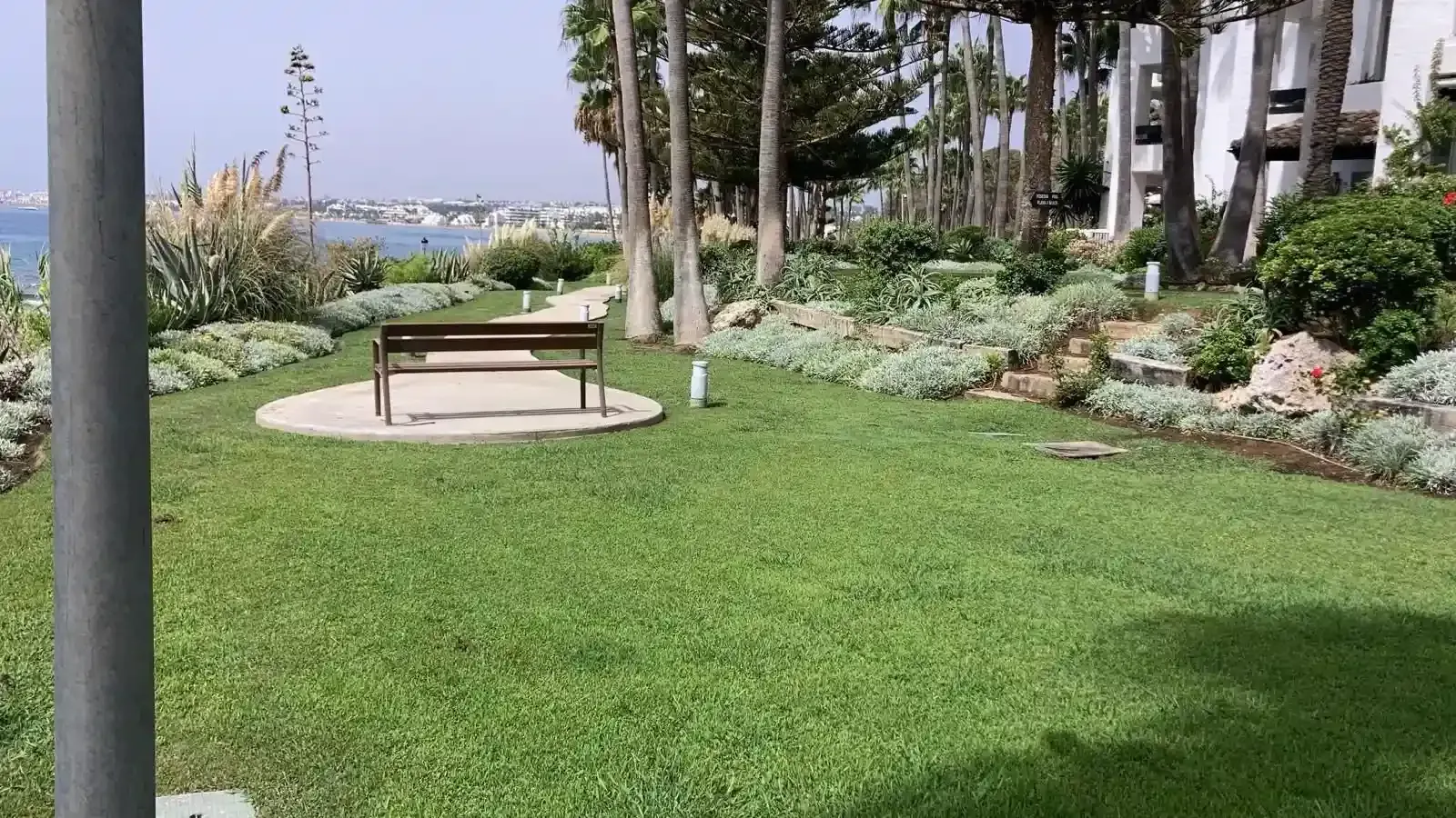 Área de parque con césped, un banco, jardines y vista al agua y a los edificios a lo lejos.