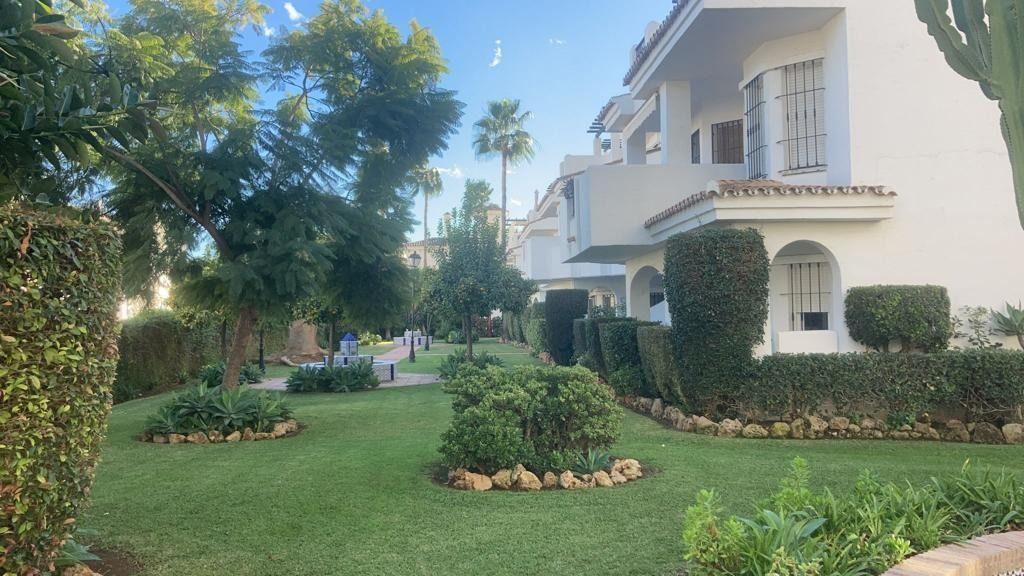 Exuberante jardín verde con setos cuidados y árboles frente a edificios blancos con puertas arqueadas.