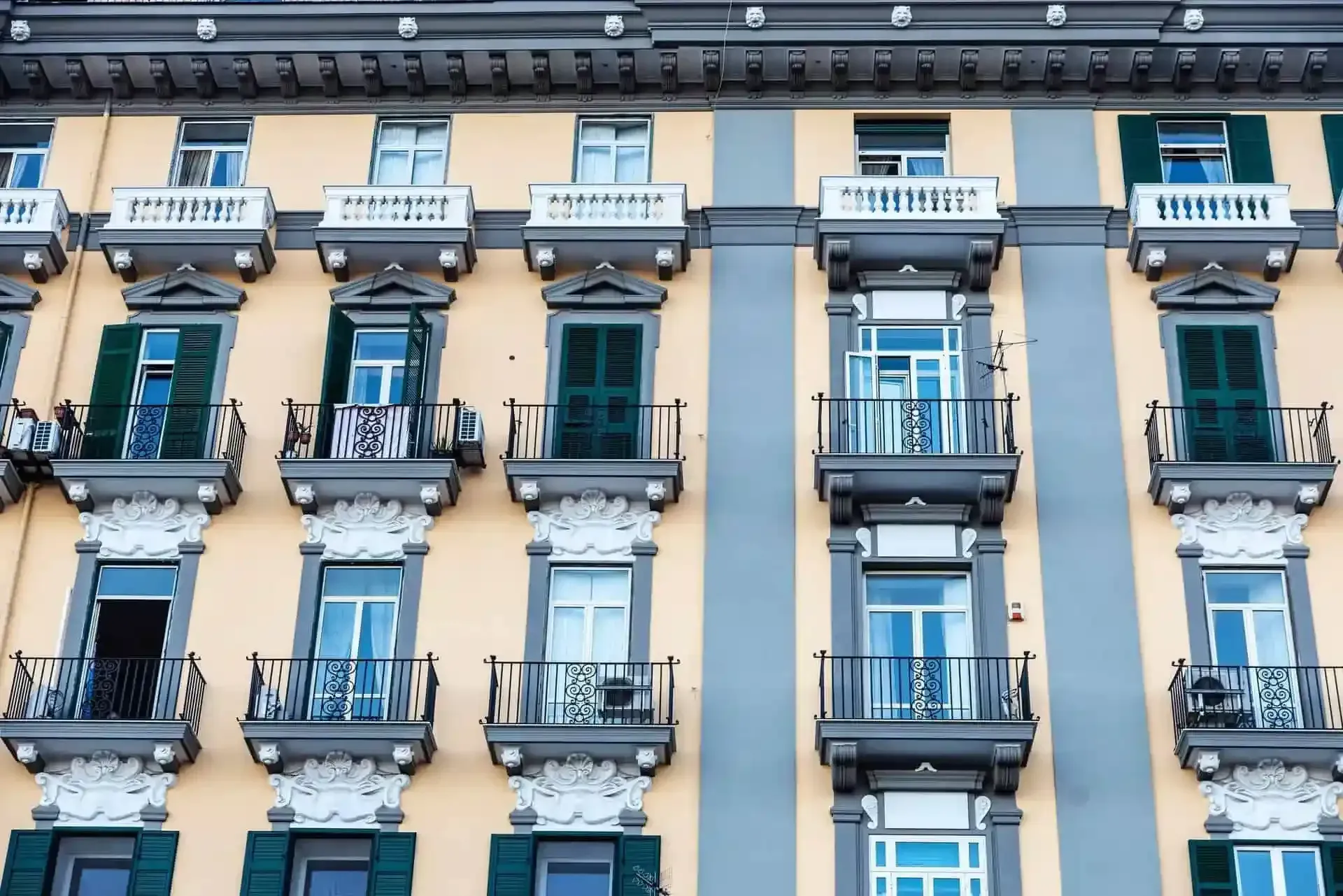 Fachada de edificio con ventanas repetidas, balcones y contraventanas verdes, pintadas en beige y gris.
