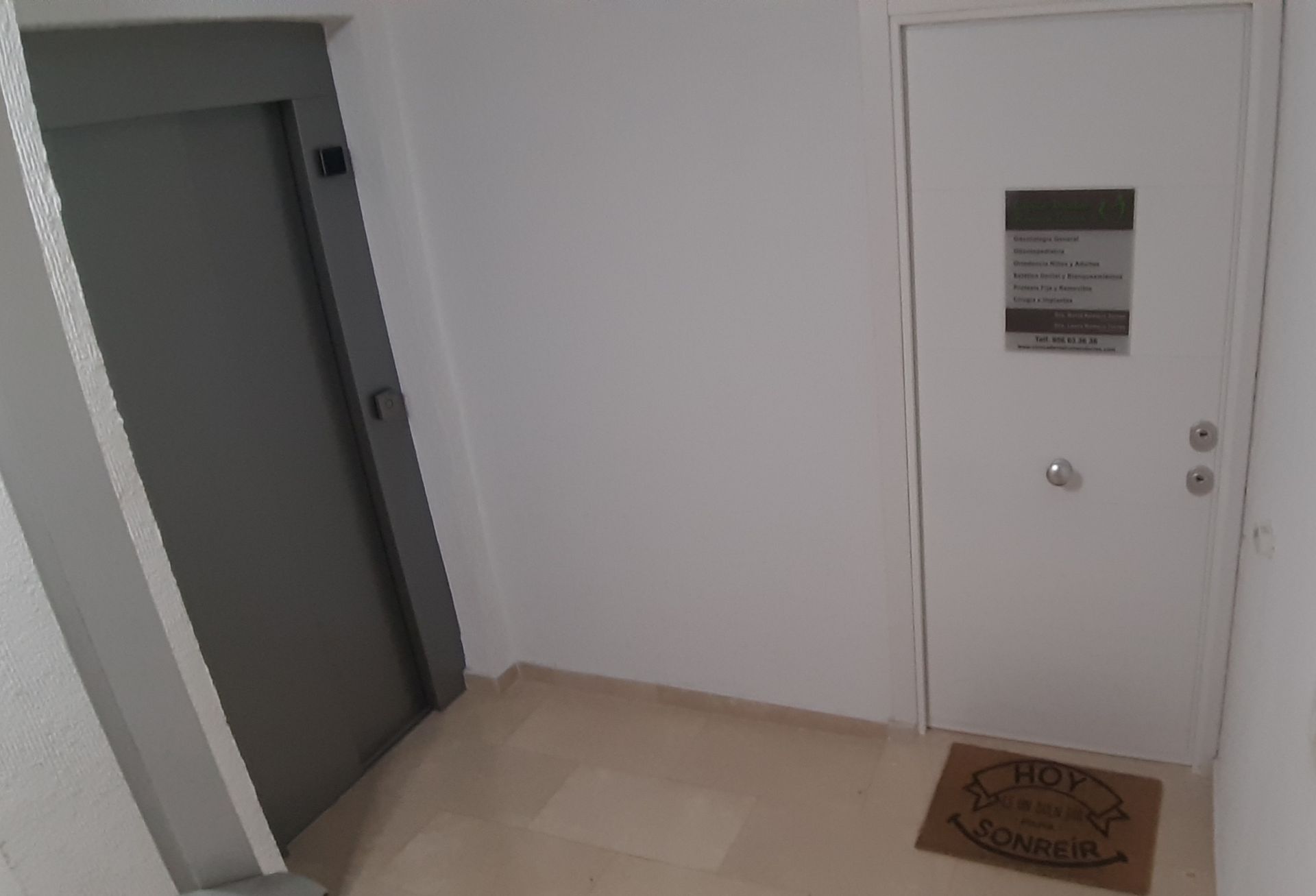 Pasillo con ascensor gris, puerta blanca y felpudo.