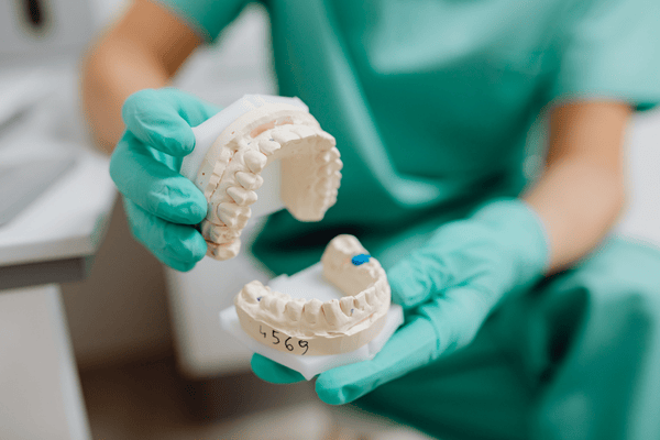 Dentista con guantes verde azulado sosteniendo moldes dentales; moldes blancos, uniforme verde, entorno clínico.