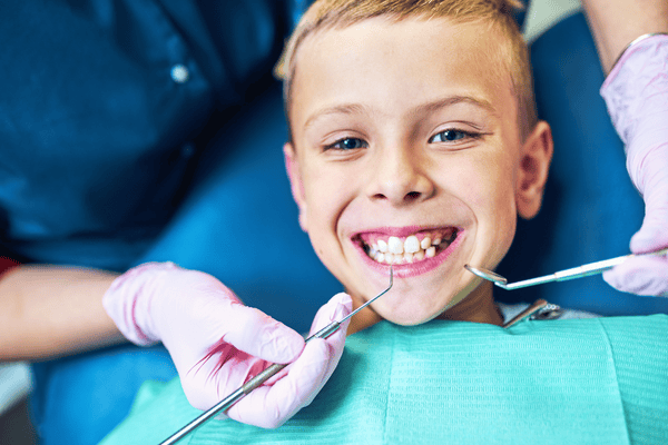 Un niño sonríe al dentista durante una revisión. El dentista, con guantes, sostiene las herramientas; el entorno es una clínica dental.