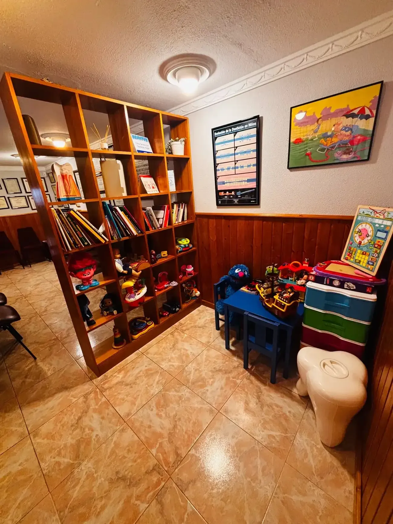 Estantería de madera llena de libros, juguetes y otros artículos en una habitación con suelo de baldosas.