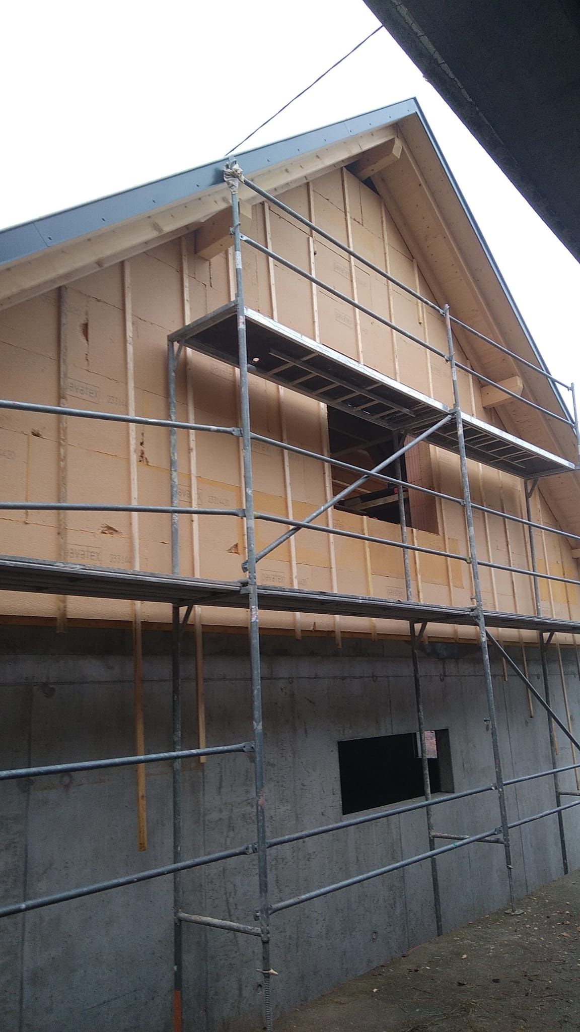 Chantier : bâtiment inachevé avec une ossature en bois et des échafaudages adossés à un mur en béton.